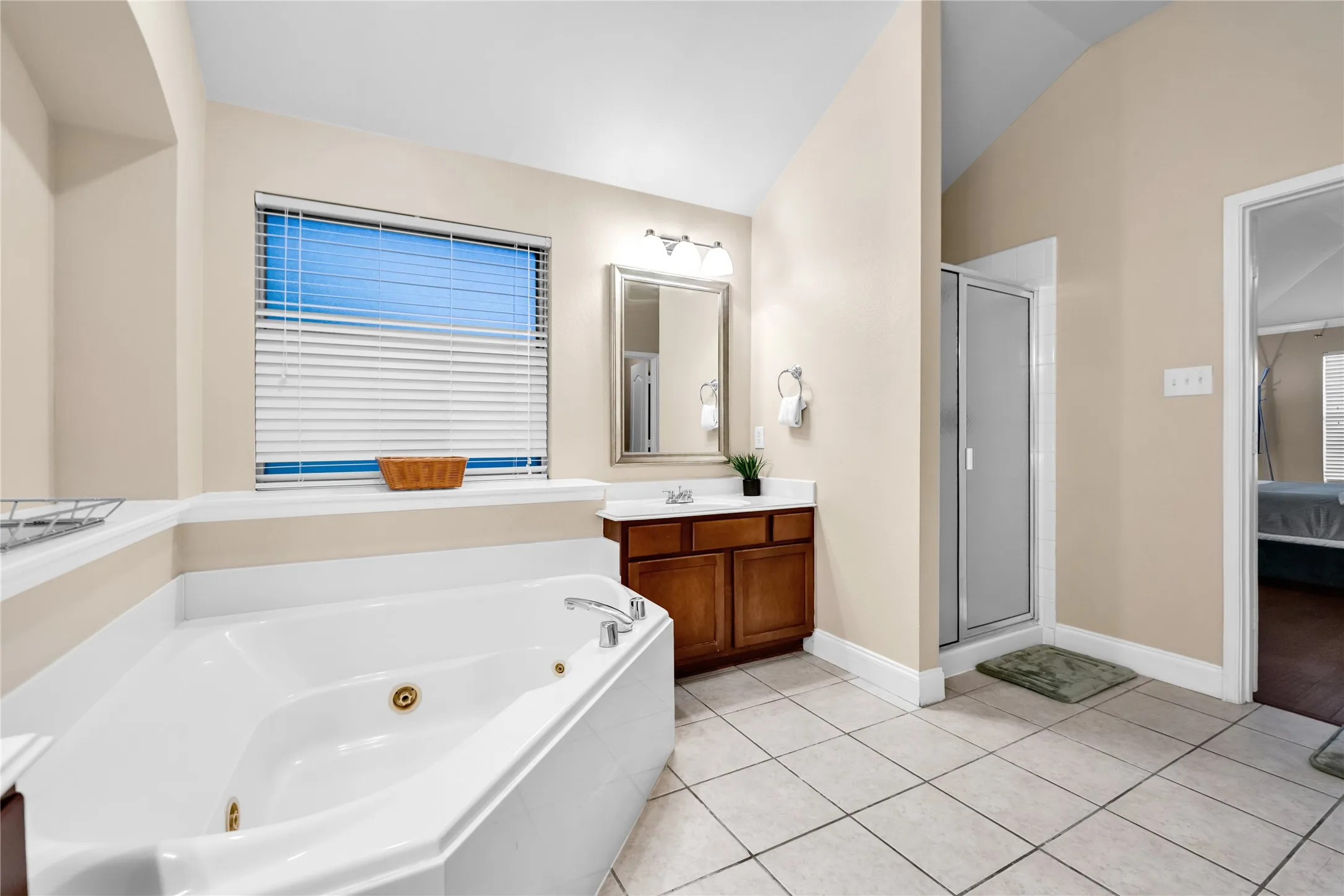 Corner jetted tub, tile flooring, wood-finish vanity, framed mirror, and a standing shower with a frosted glass door