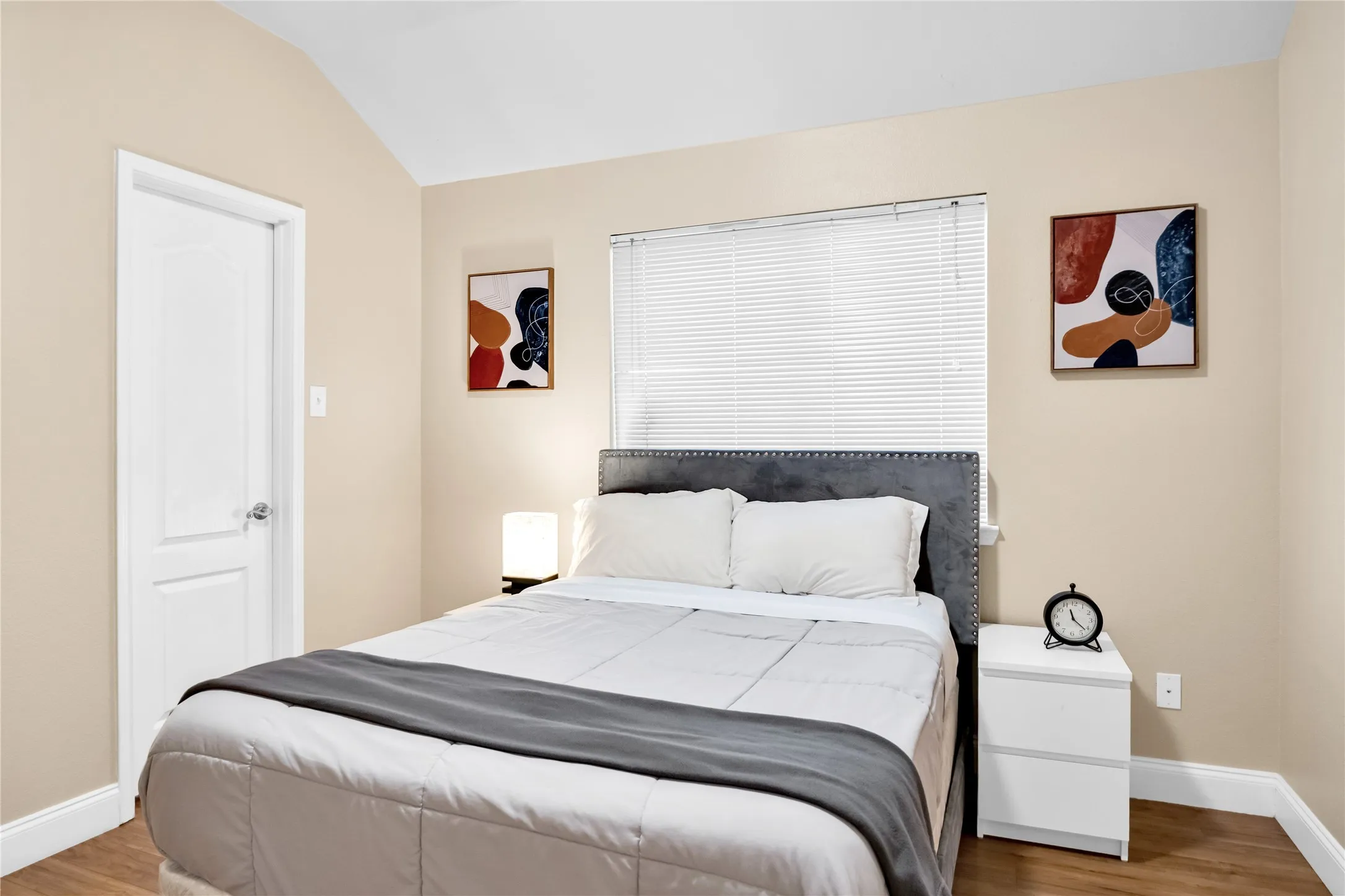 Wood-finish flooring, vaulted ceiling, white paneled door, window with blinds, and light-toned walls