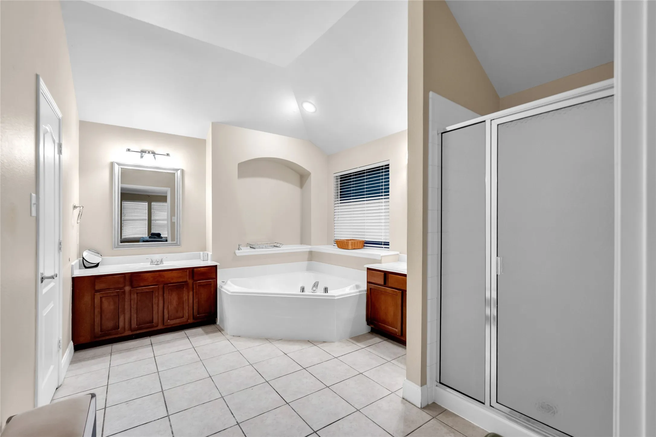 Spacious bathroom featuring a corner soaking tub, separate frosted glass shower, dual wood-finish vanity with white countertops, and neutral tile flooring
