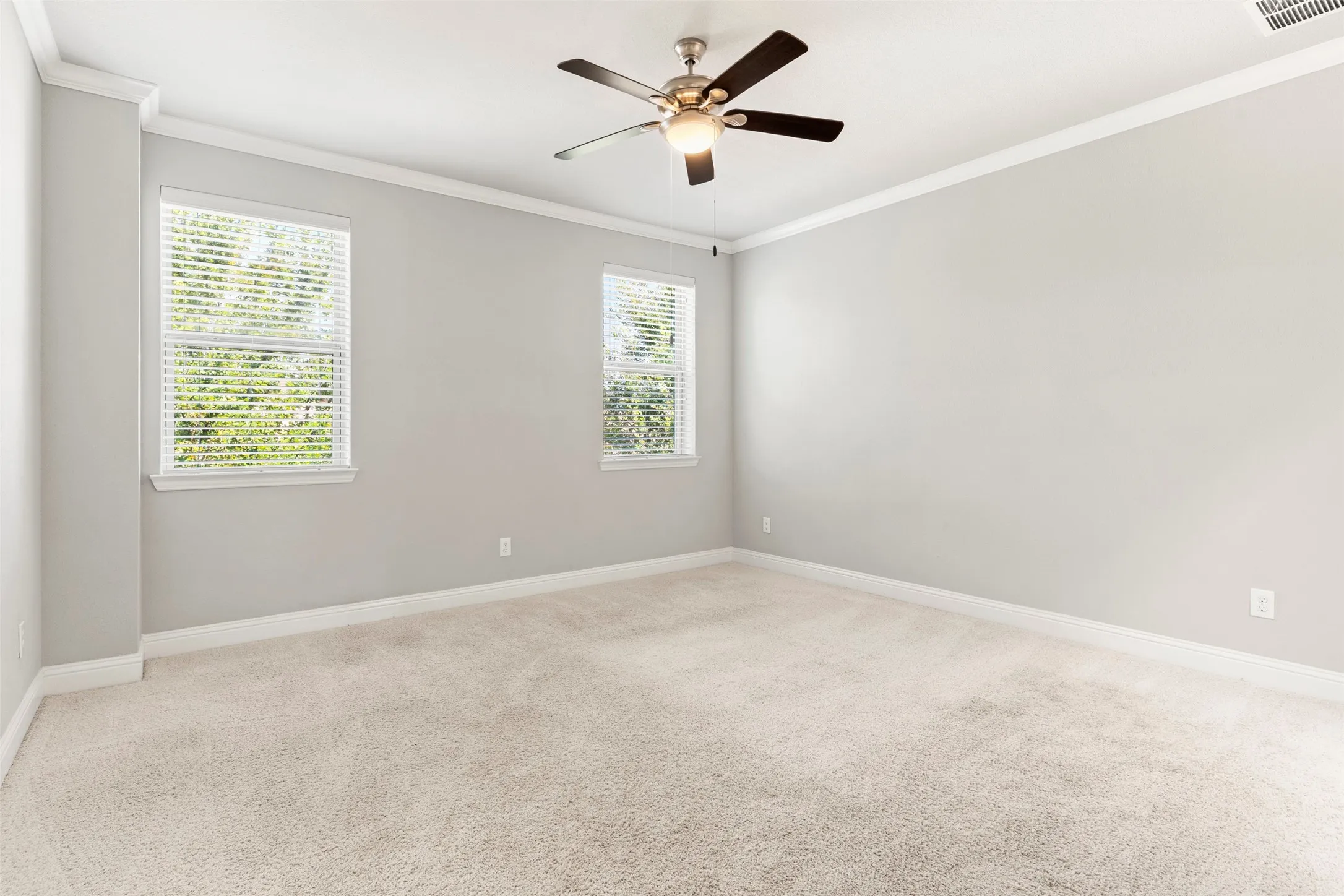 Spacious room featuring neutral carpeting, two windows with blinds, a ceiling fan with integrated lighting, and crown molding