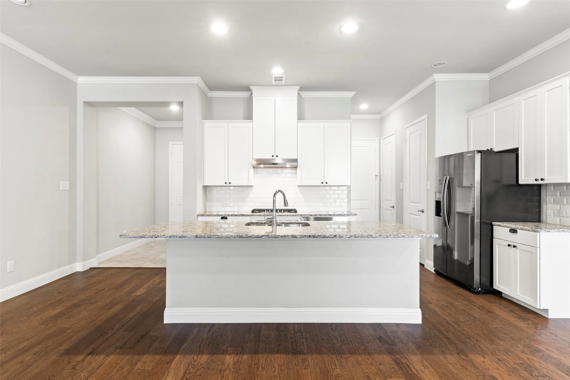 Modern kitchen featuring a large center island with granite countertops, stainless steel appliances including a French-door refrigerator, white cabinetry with ample storage, subway tile backsplash, and warm wood-finish flooring throughout the open living space
