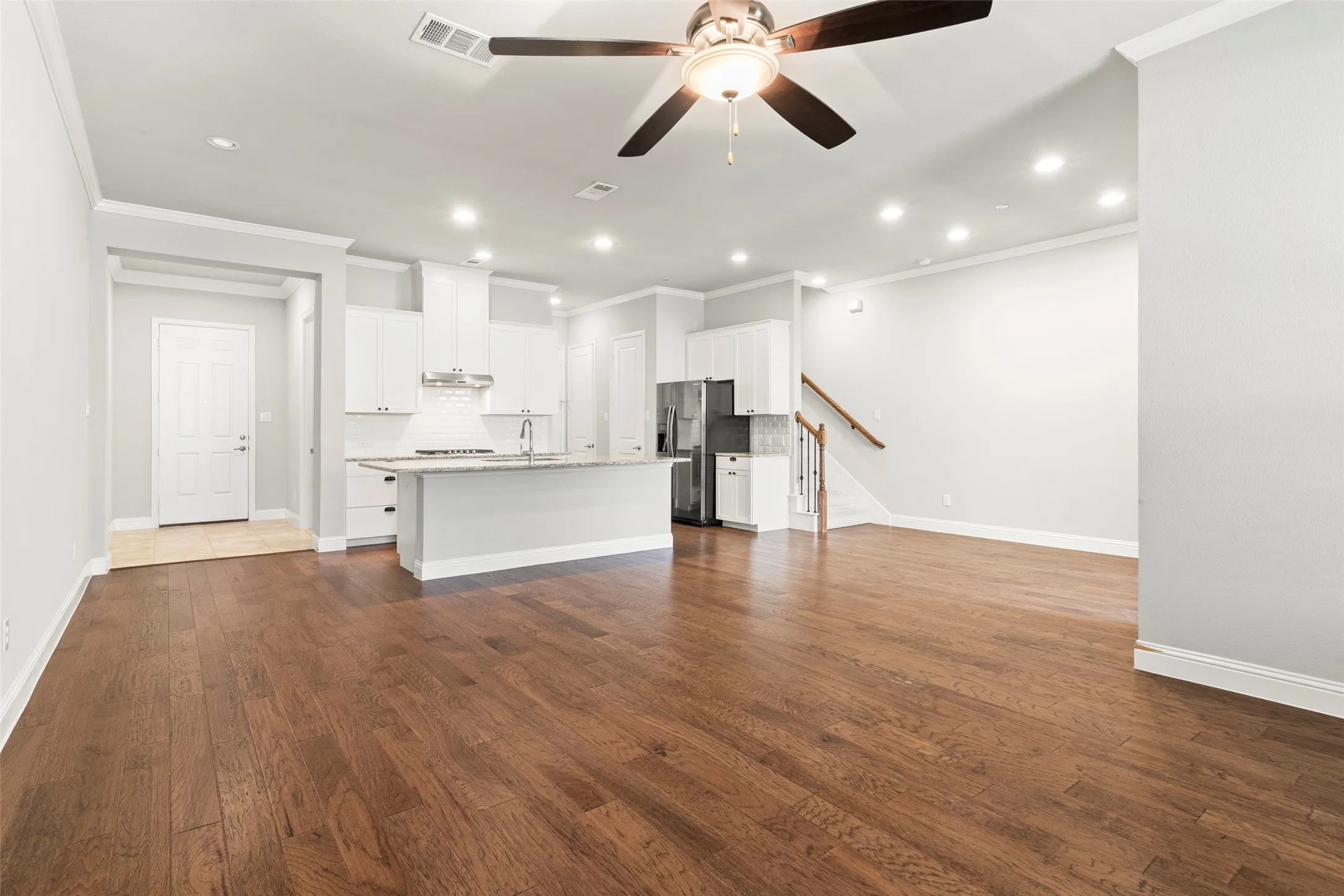 Open-concept living space featuring hardwood flooring throughout, a white cabinetry kitchen with stainless steel appliances and an island, recessed lighting, a ceiling fan with integrated light fixture, and a curved staircase with metal railing leading to the upper level