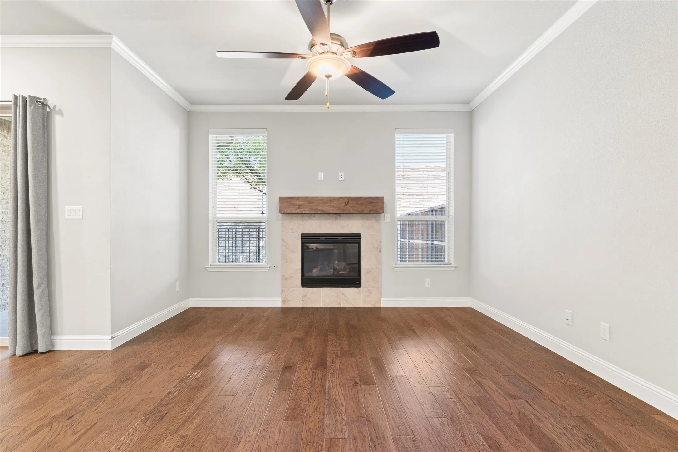 Spacious living room featuring warm wood-finish flooring, a ceiling fan with integrated lighting, and a centered fireplace with a wood mantel and stone surround