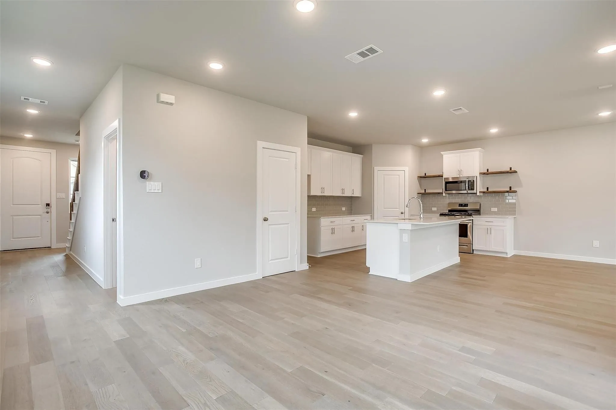 Light wood-finish flooring throughout, recessed ceiling lighting, kitchen island with built-in sink and faucet, stainless steel appliances, and white cabinetry with open shelving