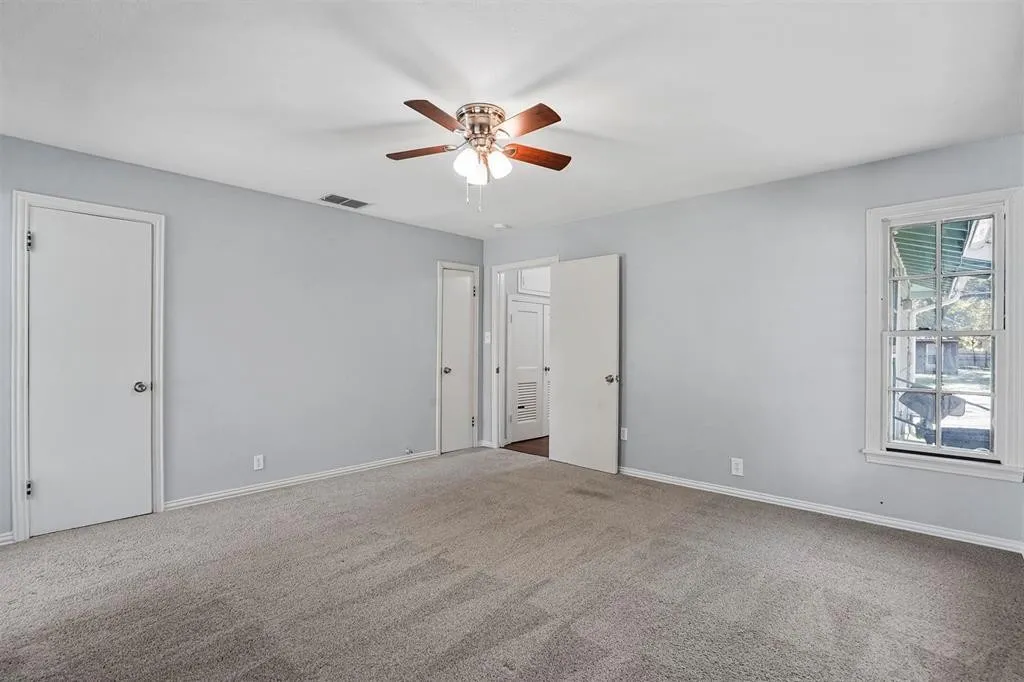 Spacious room featuring light gray wall paint, gray carpet flooring, a ceiling fan with wood-finish blades, and a white framed window