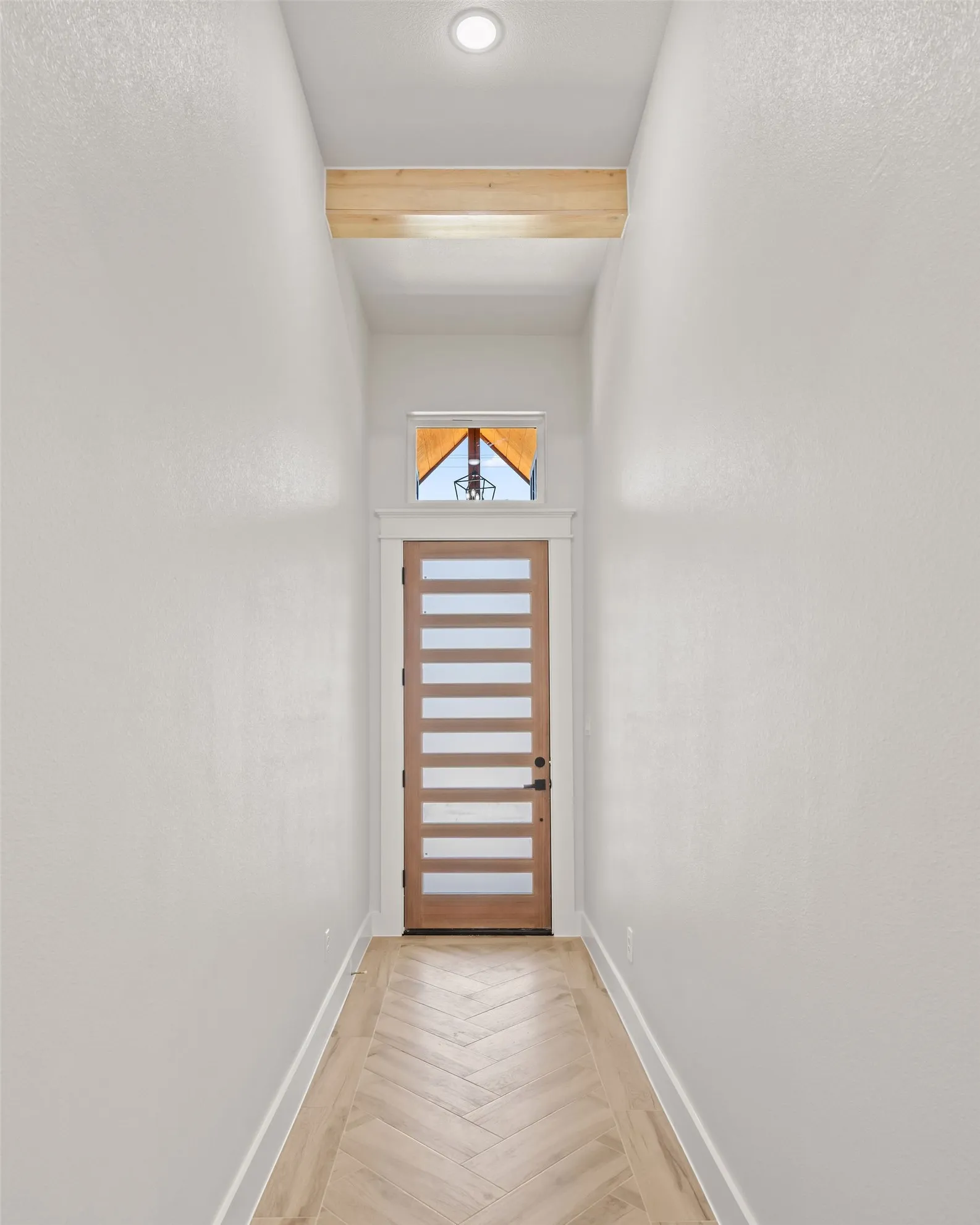 Herringbone pattern wood-finish flooring, recessed ceiling lighting, light wood door with horizontal glass panels, and a transom window with exposed beam