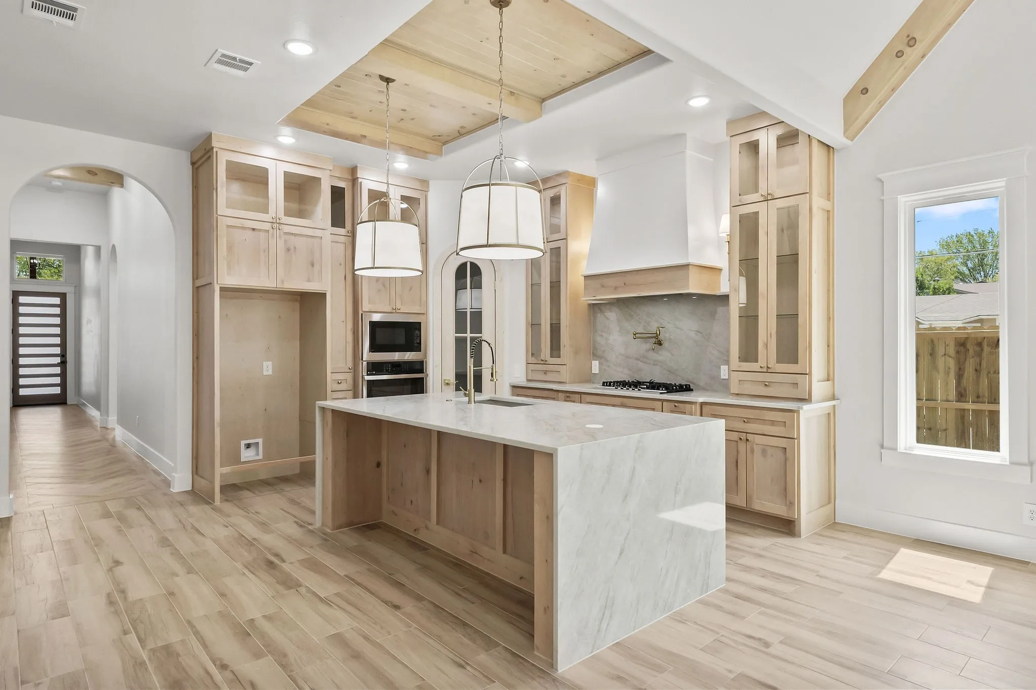 Kitchen featuring wood-finish flooring, a large island with a stone countertop, light wood cabinetry with glass fronts, a decorative range hood, and integrated double ovens