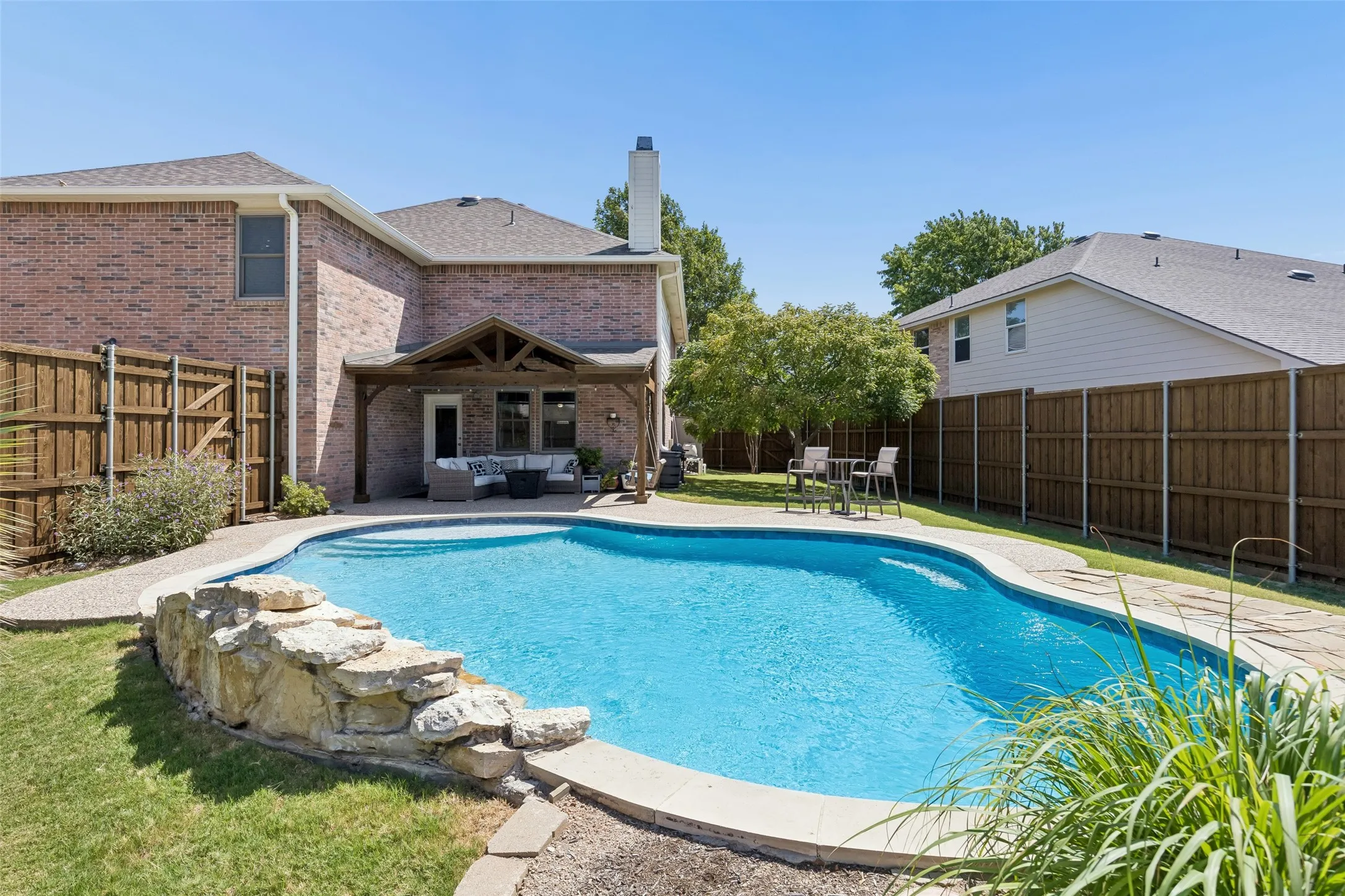 A Pool with a Rock Waterfall – The freeform swimming pool features a rock waterfall that adds movement and character to the backyard, complemented by landscaping and a broad surrounding deck.