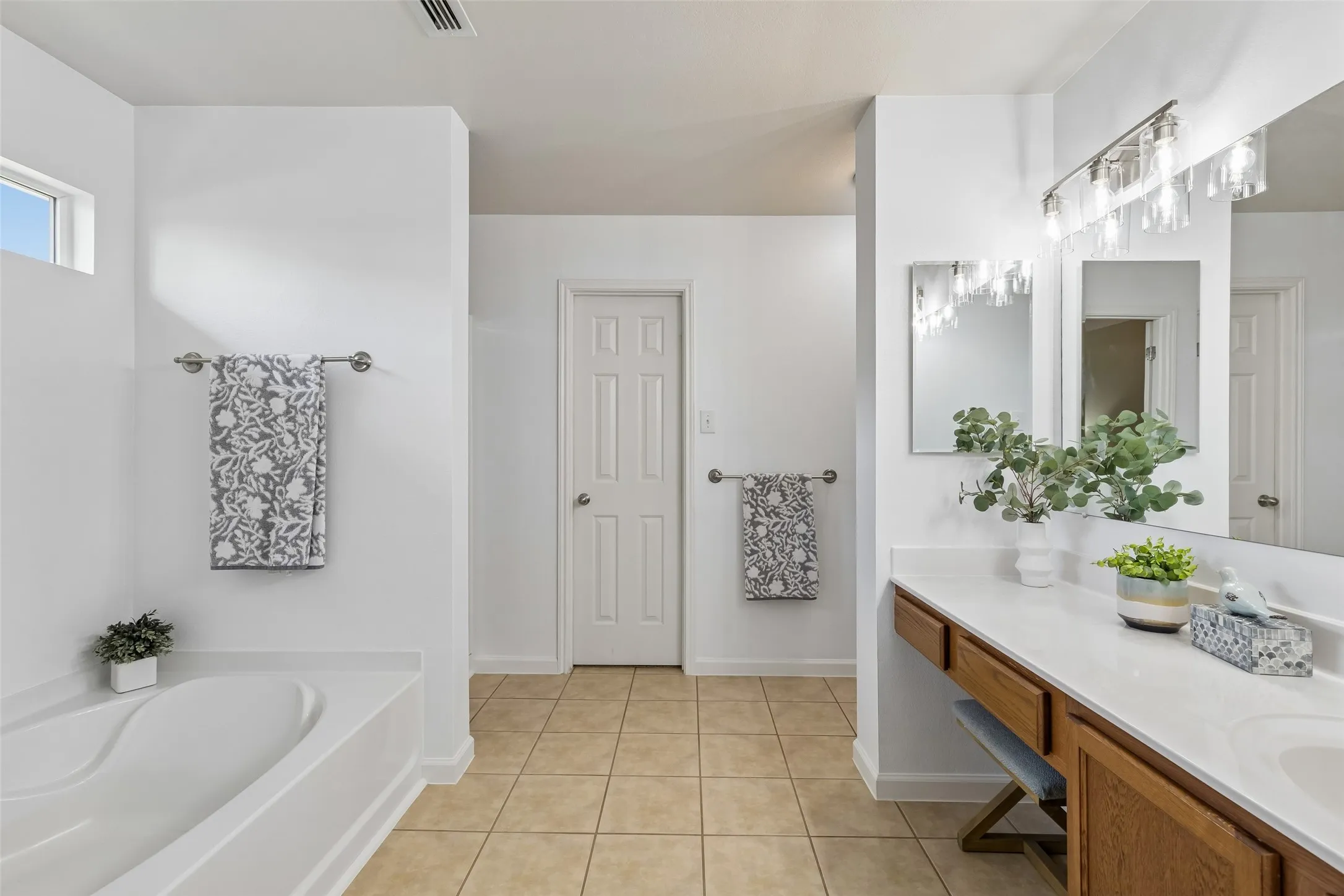 A Spacious Primary Bath – A long vanity with a dedicated dressing area, updated lighting, tiled flooring, and a separate tub create a functional bath with plenty of room to move comfortably through the space.