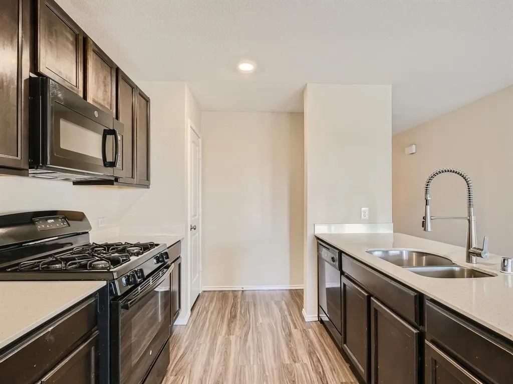 Kitchen featuring wood-finish flooring, dark wood cabinetry, light countertops, stainless steel double basin sink with gooseneck faucet, and a built-in microwave over a gas range