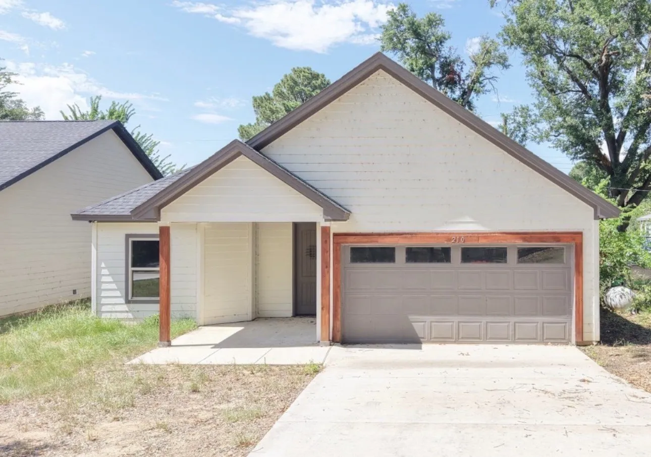 Gabled roofline with dark shingles, light siding exterior, integrated garage with panel doors, covered entryway with wooden support beams, and a concrete driveway