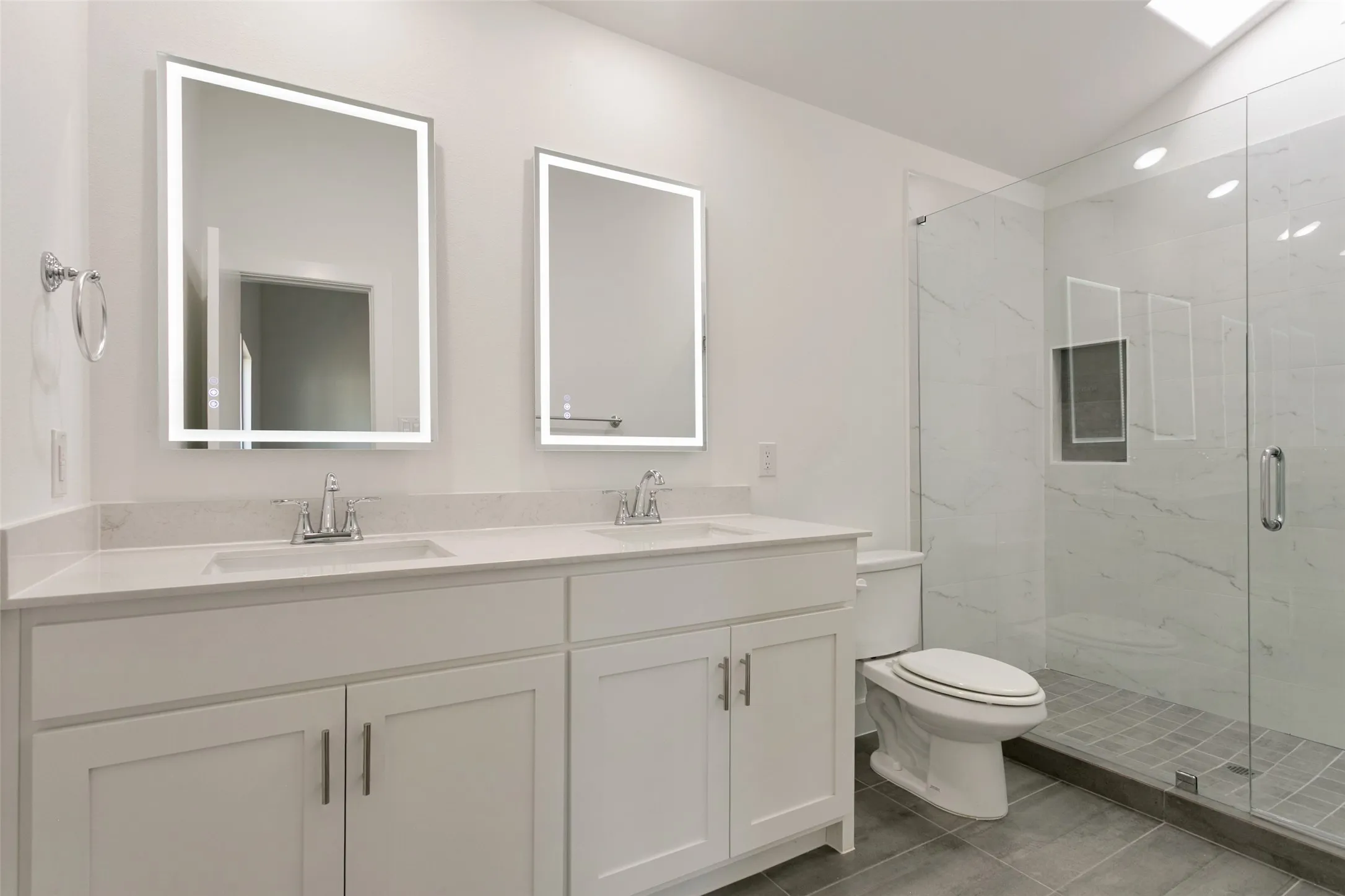 Dual vanity with integrated sinks, illuminated mirrors, glass-enclosed shower, skylight, and large format floor tile