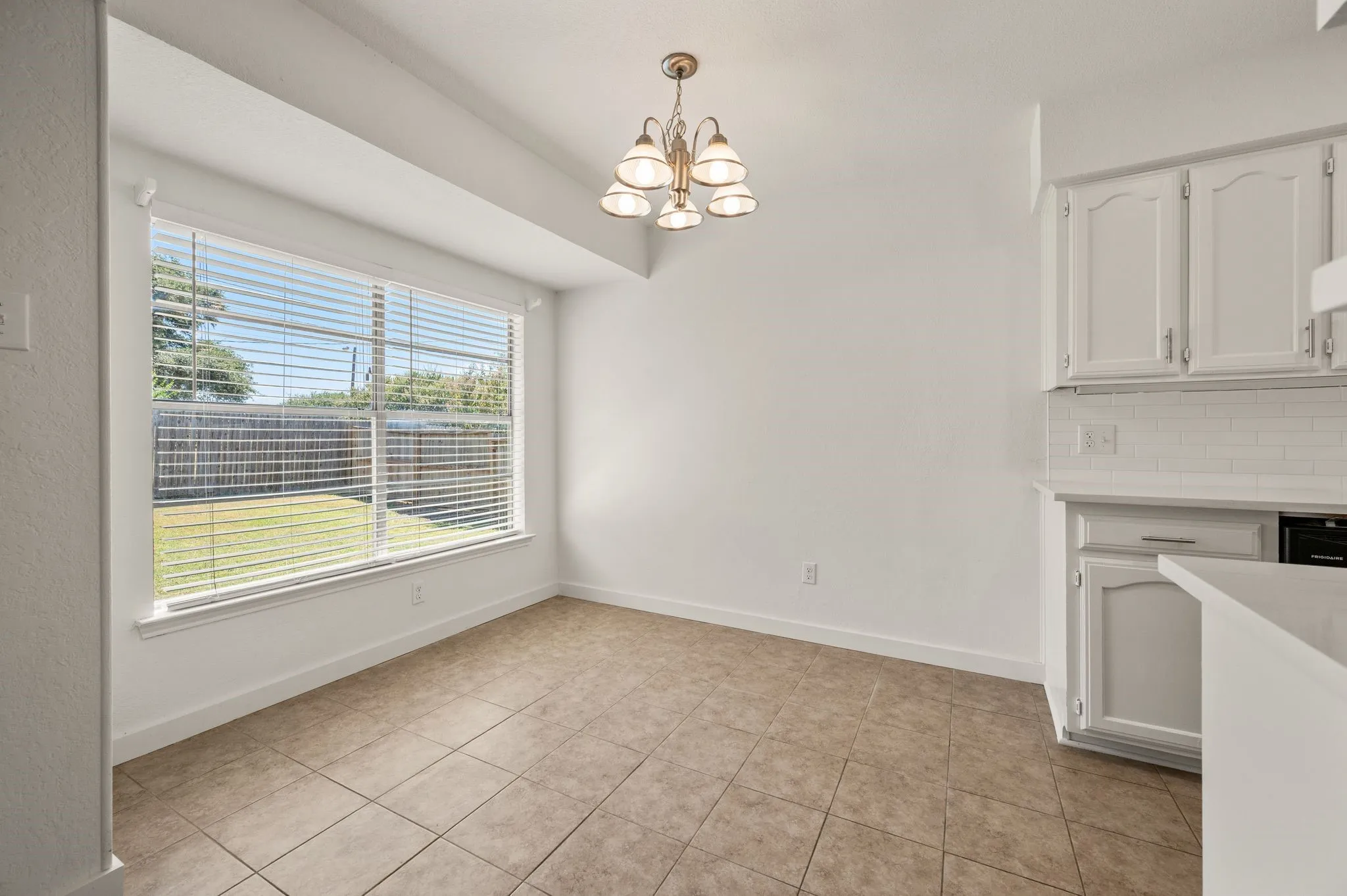 Bright, light-filled kitchen and dining area featuring large picture window with natural outdoor vistas, ceramic tile flooring, white cabinetry with integrated appliances, and brushed gold pendant lighting