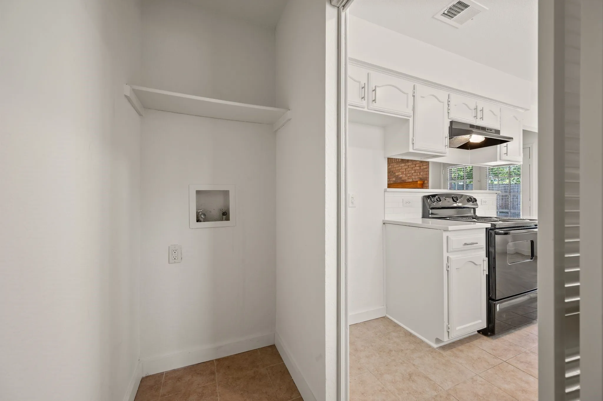 Integrated laundry alcove with convenient shelving and hookups, adjacent to a kitchen featuring white cabinetry, a dark range, and a tile backsplash