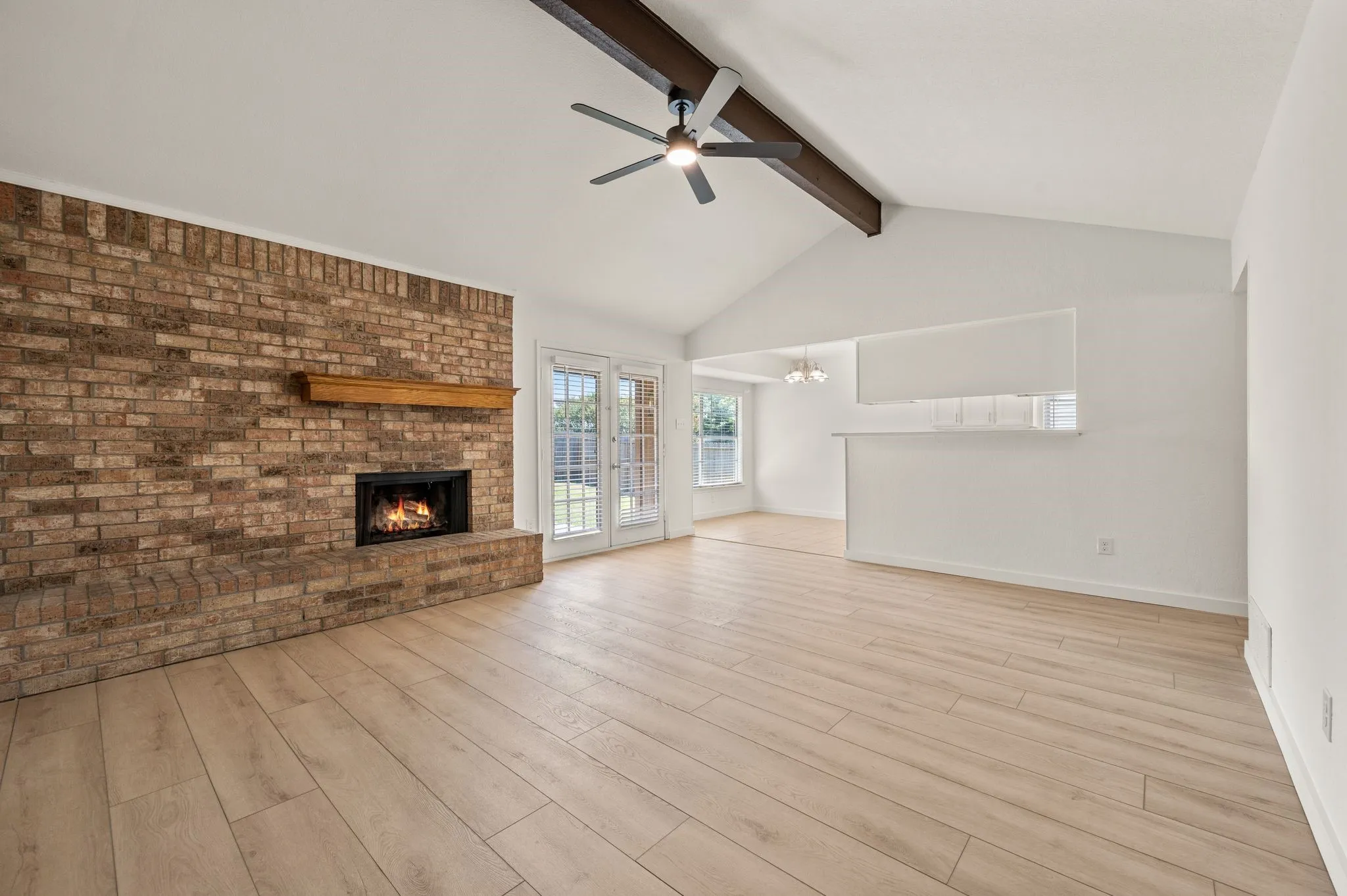 Brick fireplace with wood mantle, vaulted ceiling with exposed wood beam, ceiling fan, wood-finish flooring, and French doors