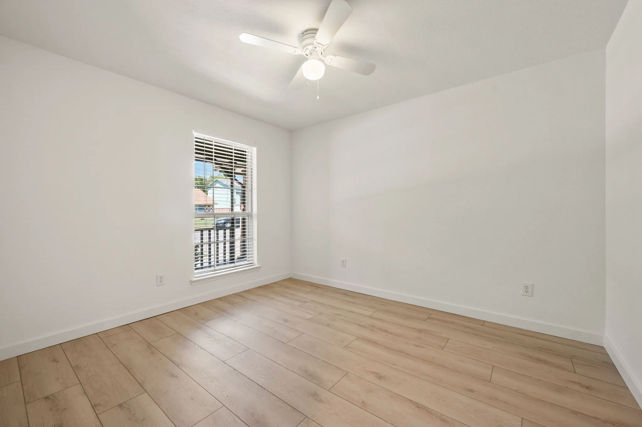 Bright and airy bedroom with wood-finish flooring, ceiling fan with integrated lighting, and a large window with plantation shutters offering natural light and ventilation