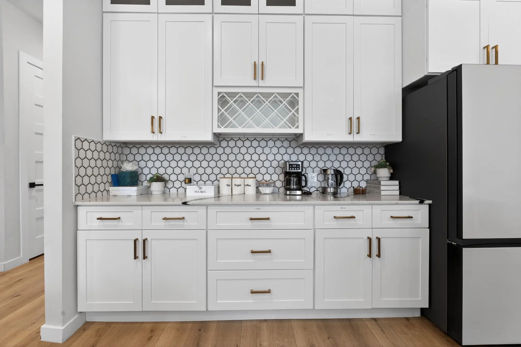 White shaker-style cabinetry with gold-tone hardware, hexagonal tile backsplash, light-toned countertops, integrated wine rack, and wood-finish flooring