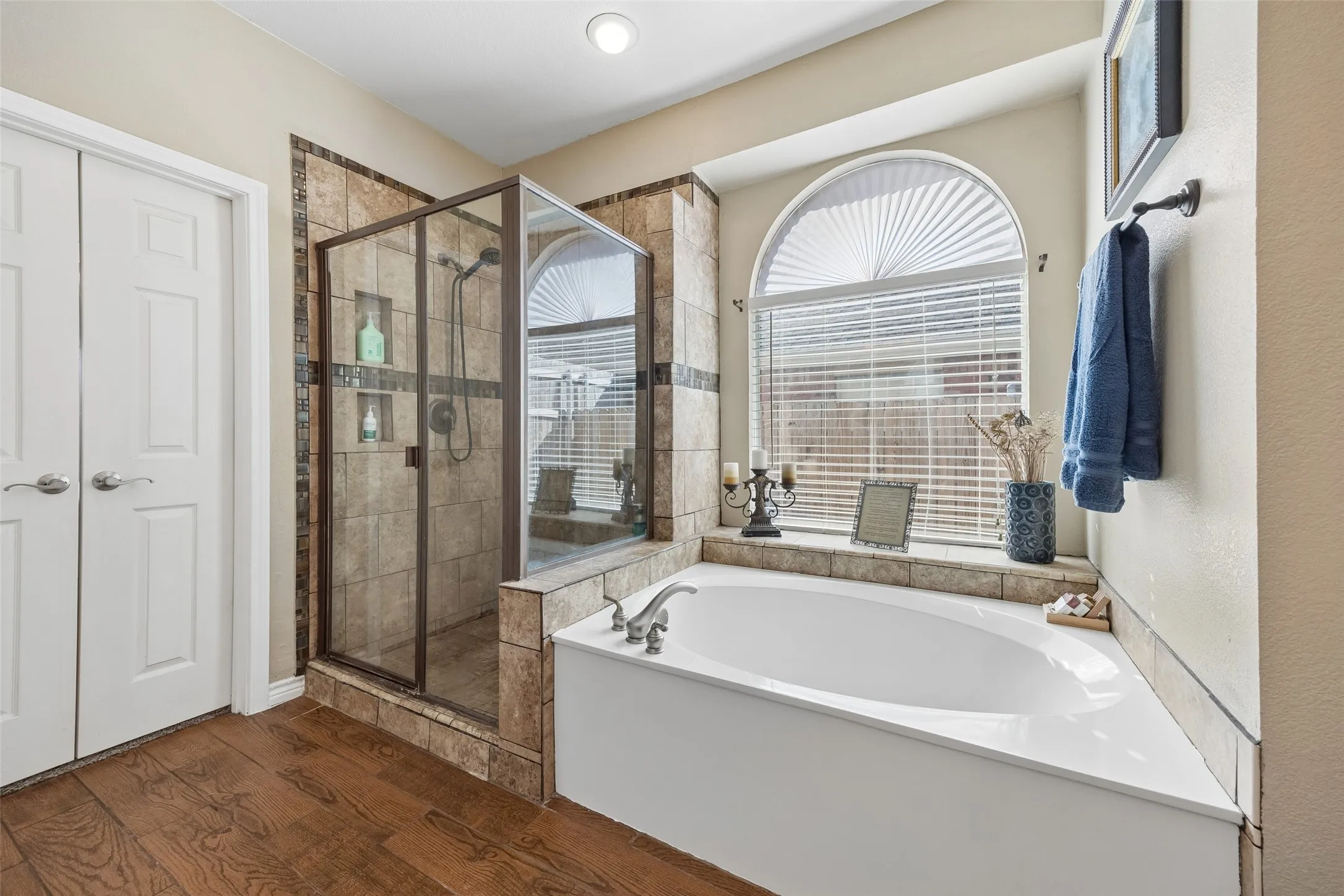 Separate Shower and Soaking Tub — The primary bath pairs a deep soaking tub beneath a large arched window with a separate tiled shower featuring a frameless-style glass enclosure, built-in niches, and decorative tile accents.