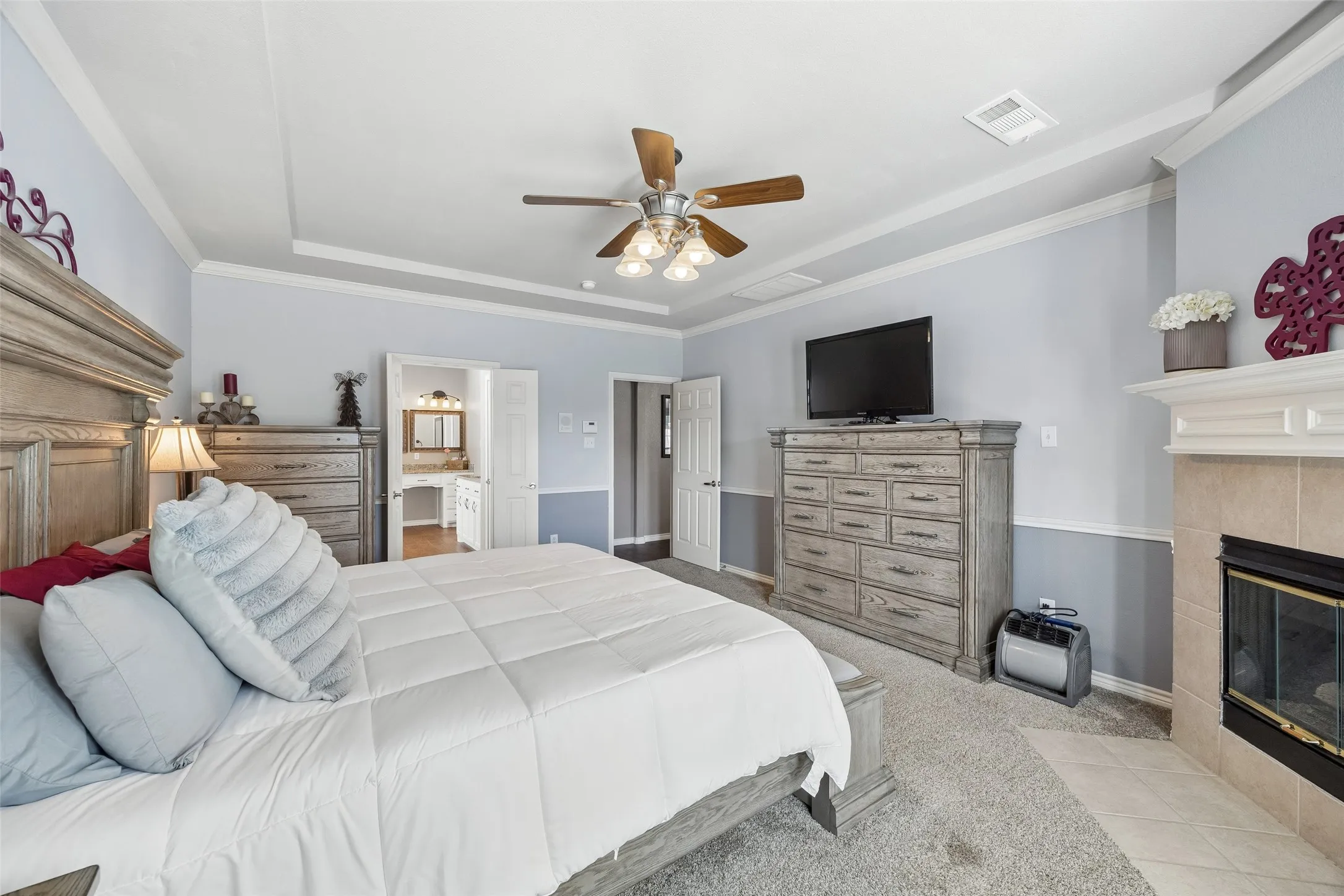 A Primary Bedroom with Extra Architectural Detail — The tray ceiling adds dimension overhead, while chair-rail molding wraps the room and the corner fireplace provides a second focal point. Direct access to the adjoining bath is visible just beyond the bedroom.