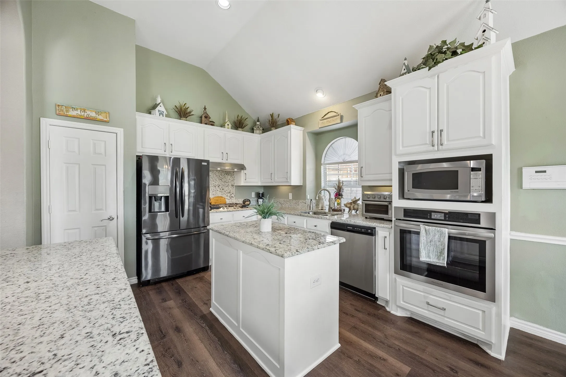 Kitchen Storage, Prep Space & Stainless Appliances — White cabinetry extends nearly to the ceiling, giving the kitchen substantial storage, while granite countertops and a center island provide multiple prep surfaces. Stainless appliances include a French-door refrigerator, dishwasher, built-in microwave, and wall oven.