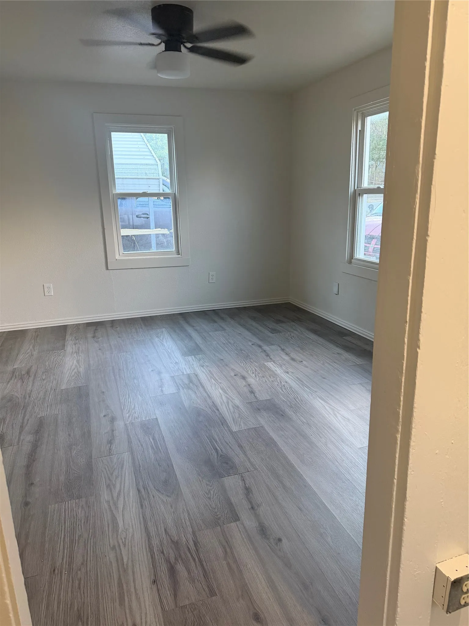 Gray wood-finish flooring with white baseboards, two windows with white trim, and a ceiling fan with integrated lighting