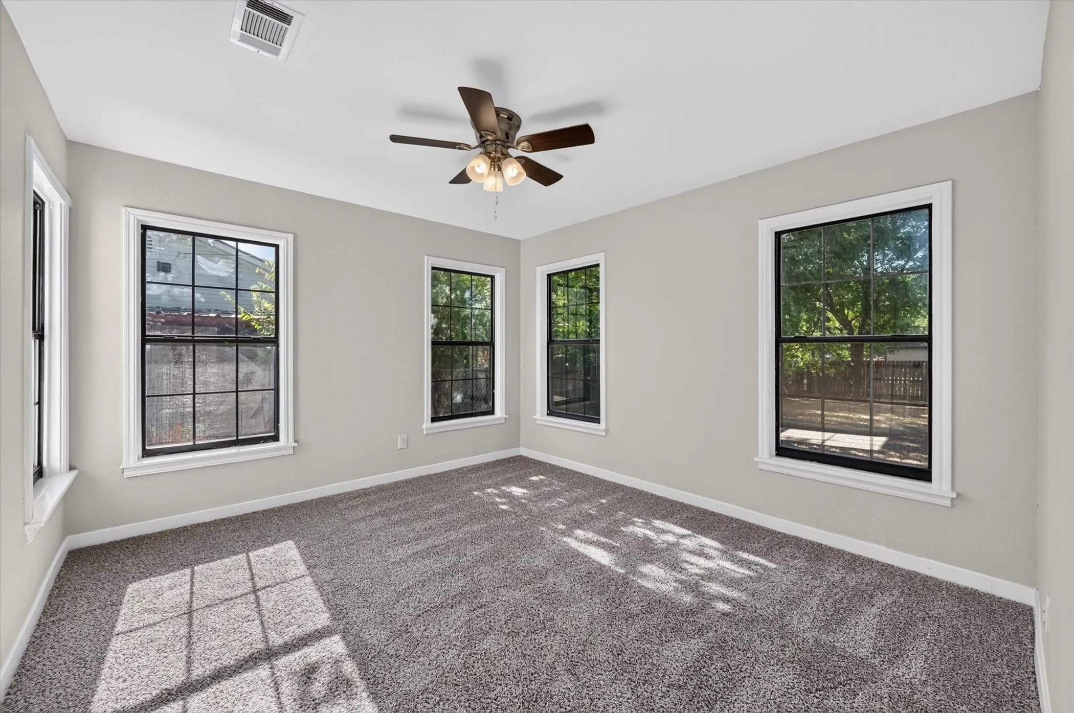 Spacious room featuring four windows with black grilles, a ceiling fan with light fixture, and neutral-toned carpeting