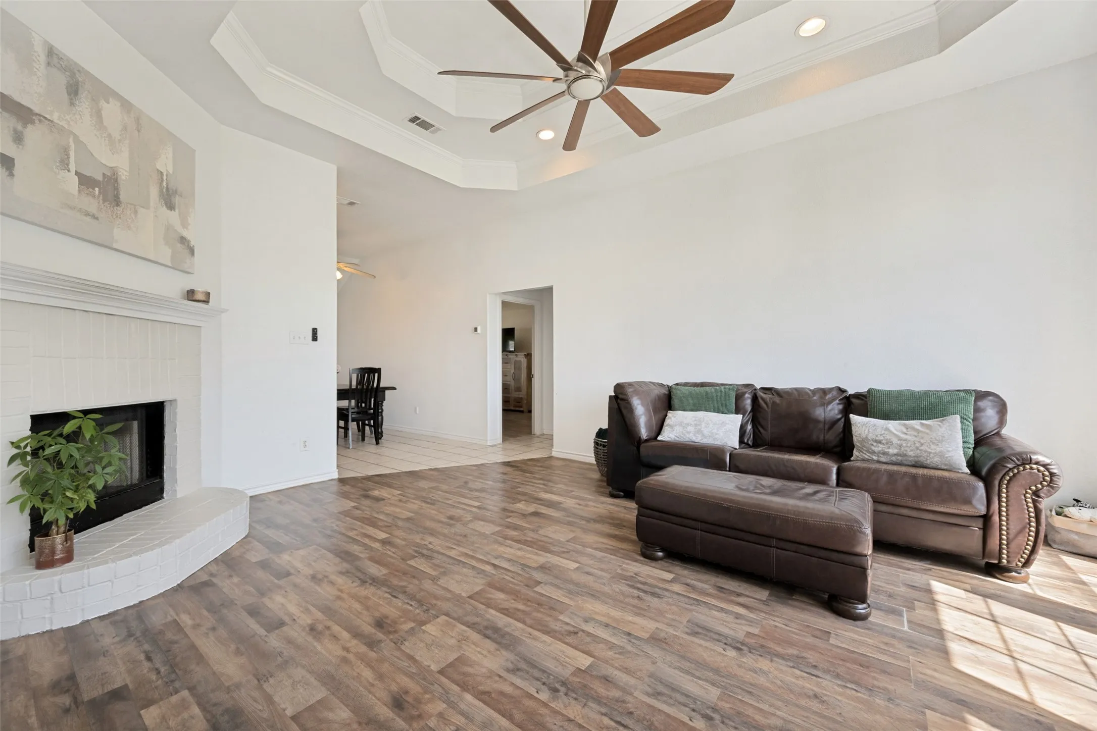 Wood-finish flooring, white brick fireplace, tray ceiling, recessed lighting, and a ceiling fan