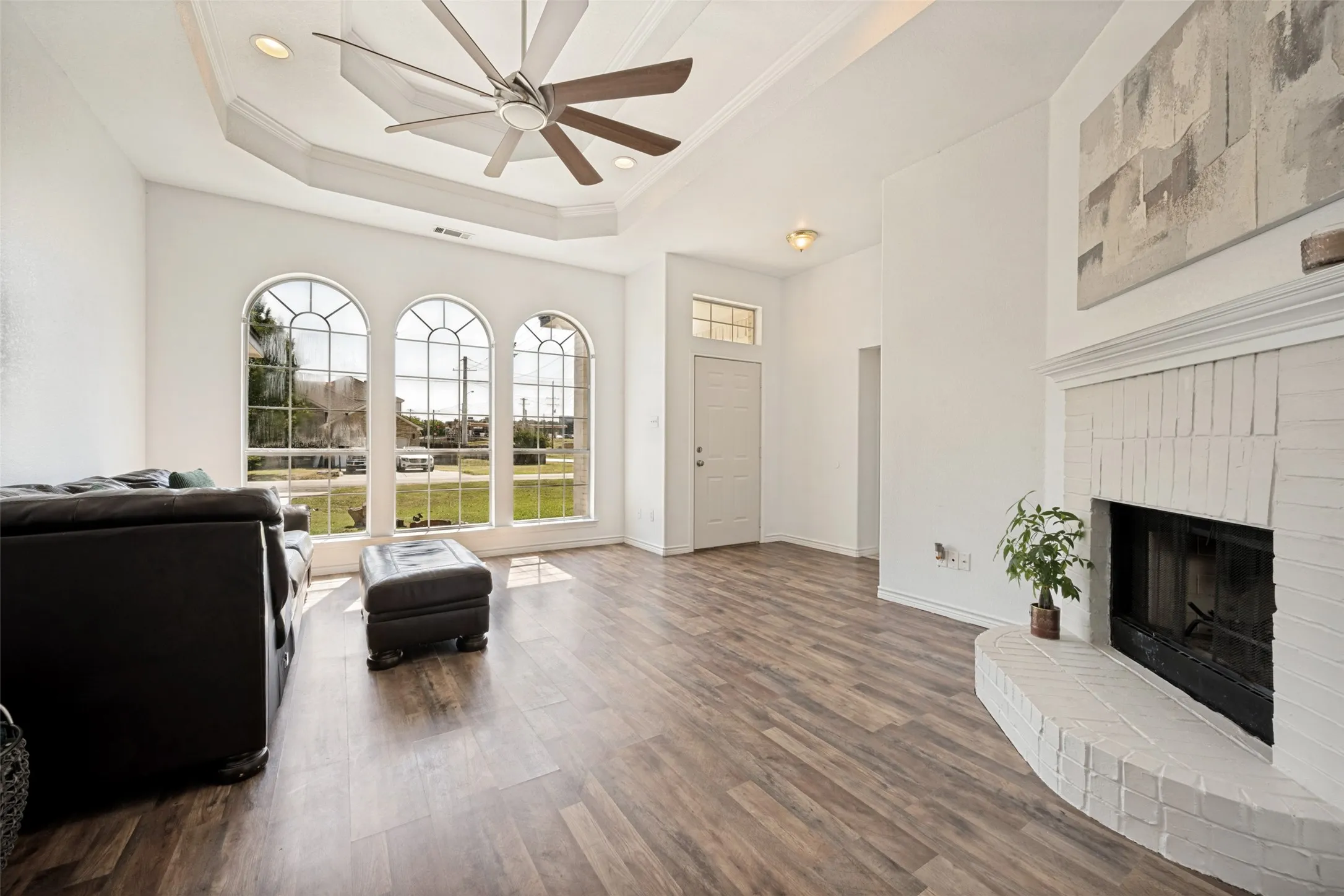 Vaulted ceiling with an integrated ceiling fan, wood-finish flooring, three arched windows with mullions, and a brick fireplace