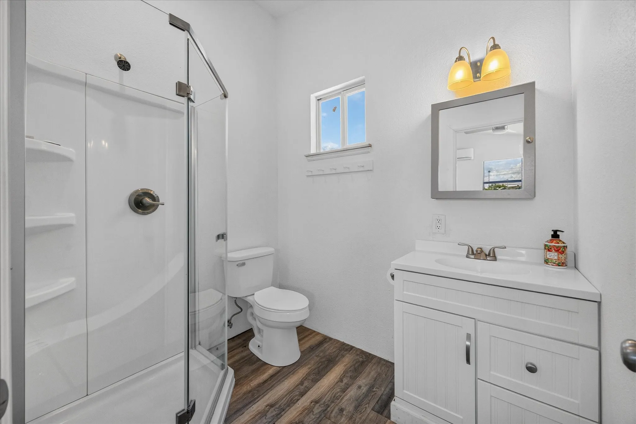 White vanity with integrated sink, wood-finish flooring, glass-enclosed shower, wall-mounted medicine cabinet, and dual-bulb light fixture
