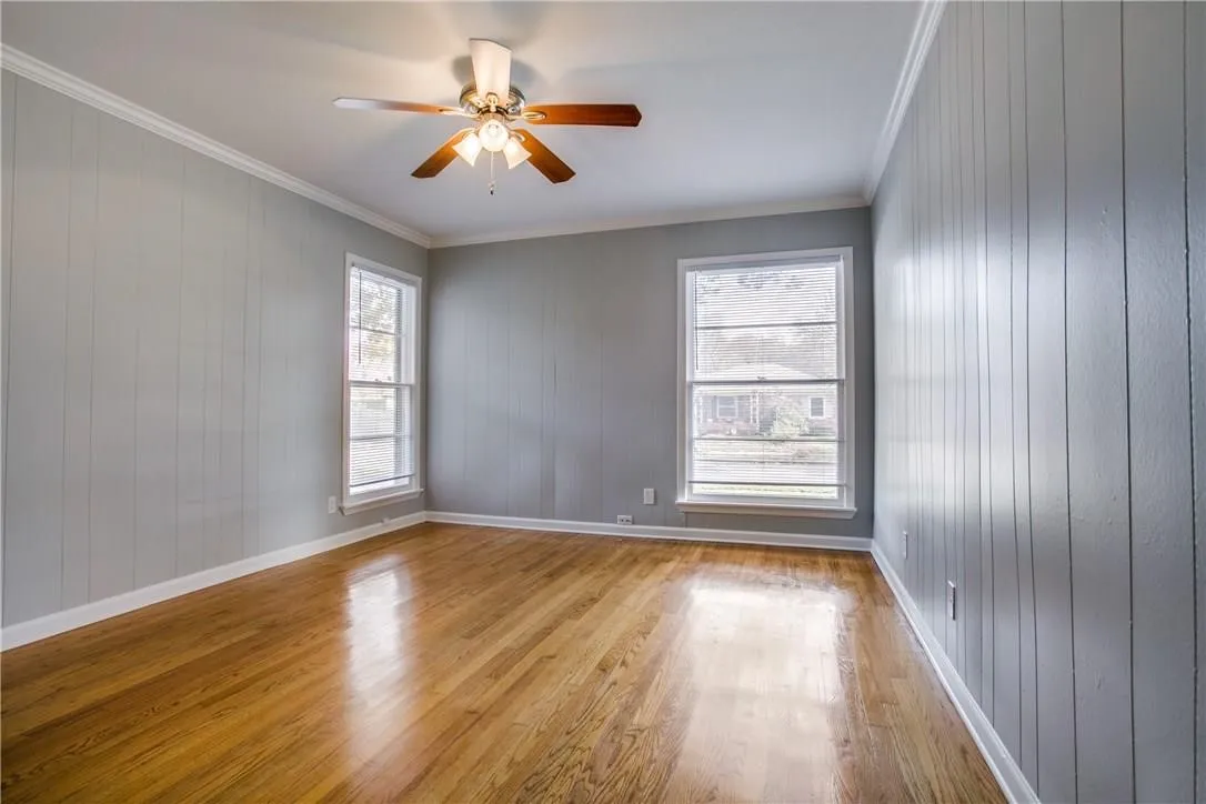 Hardwood flooring, two large windows, crown molding, vertical paneling, and a ceiling fan with integrated lighting