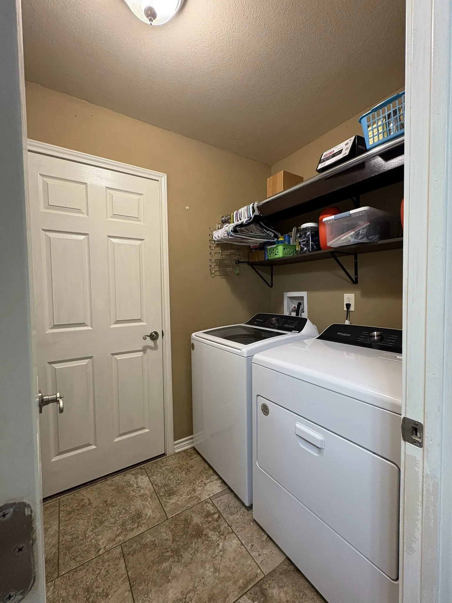 Dedicated laundry room featuring stacked white front-load washer and dryer, black countertop workspace, floating black shelving for storage, tile flooring, and recessed ceiling lighting
