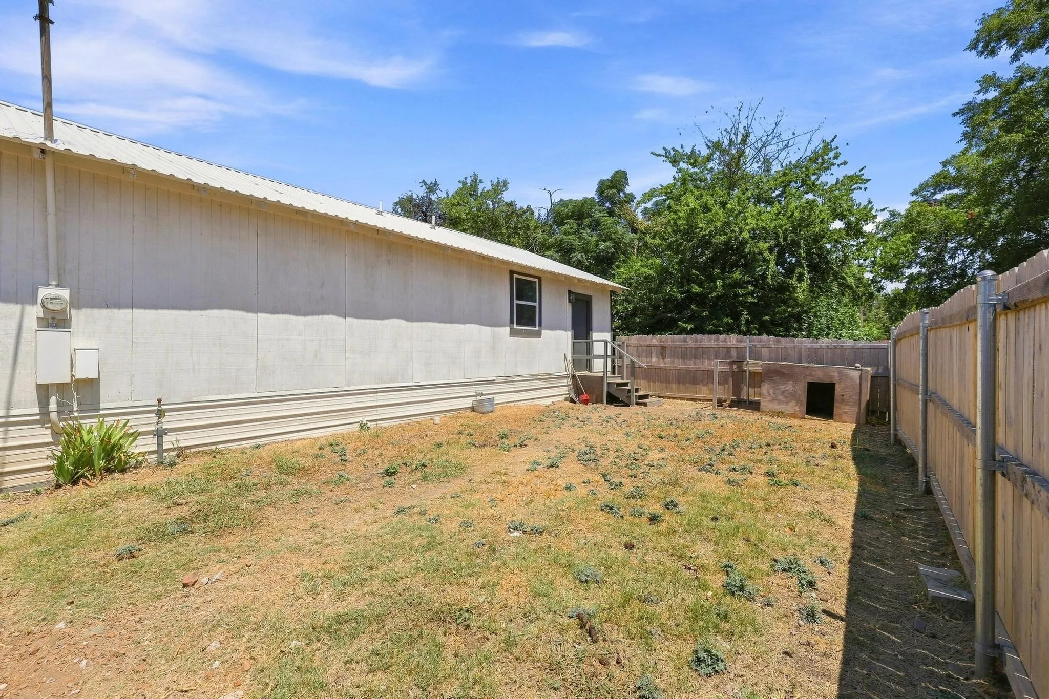 Spacious backyard with level lawn and mature tree coverage, fenced perimeter, and single-wide manufactured home featuring metal siding, covered entry, and window placement for natural light