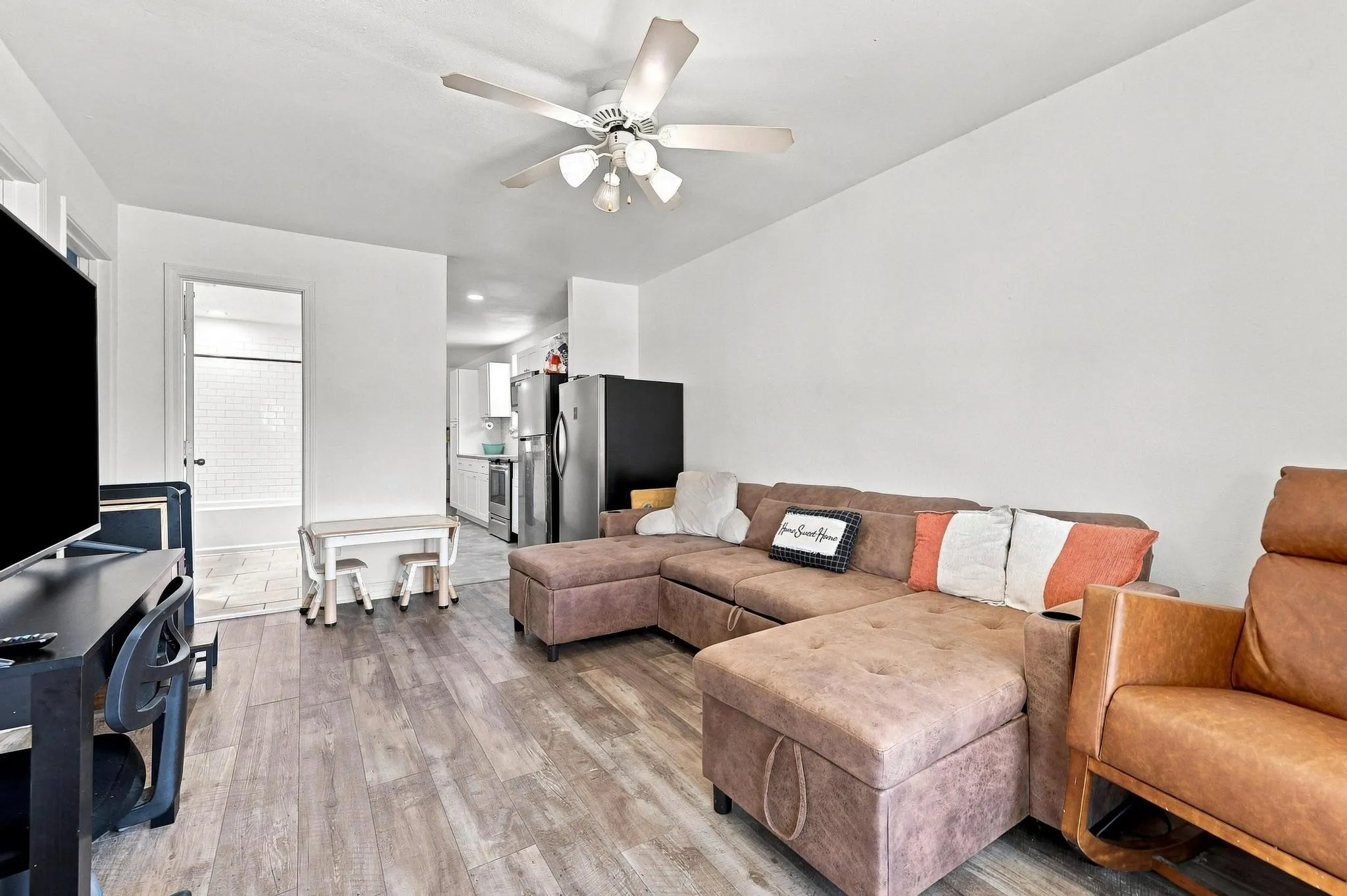 Open-concept living area featuring plush sectional seating, wood-finish flooring throughout, ceiling fan with integrated lighting, stainless steel refrigerator, and direct access to the entry foyer with natural light from front-facing windows