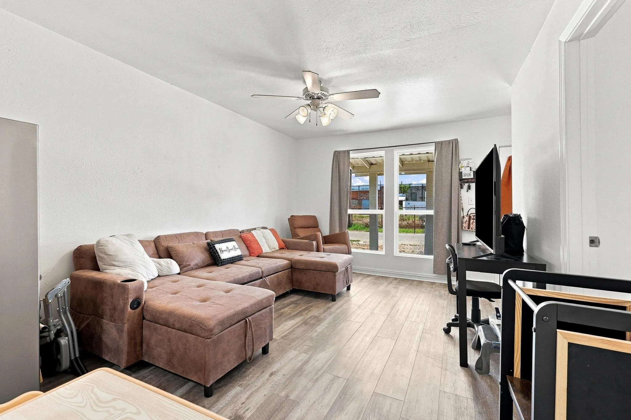 Spacious living area featuring a brown leather sectional with ottoman, wood-finish flooring throughout, ceiling fan with integrated lighting, glass sliding doors with transoms providing natural light, and a dedicated entertainment zone with built-in media console and shelving