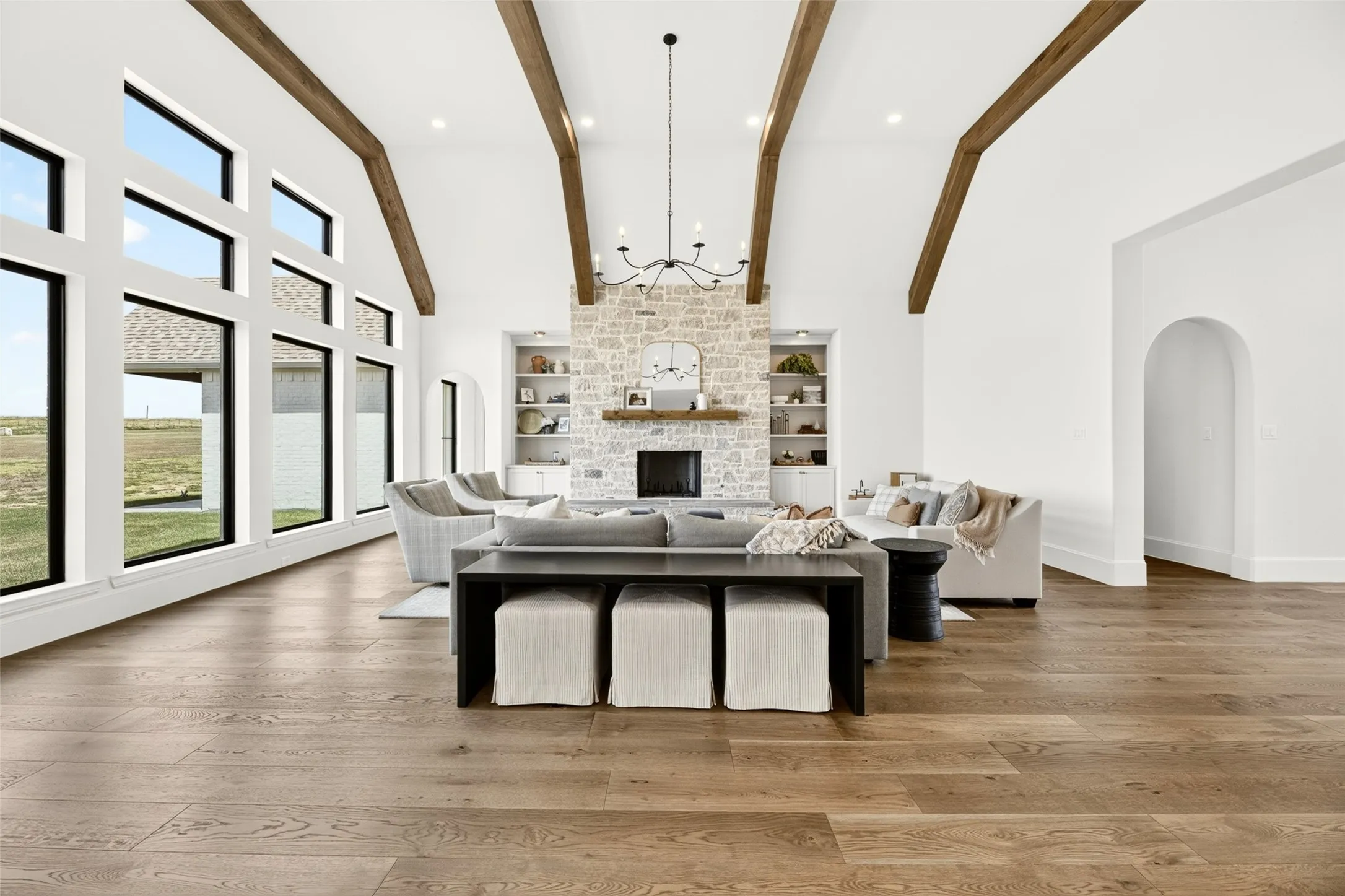 Vaulted ceiling with exposed wood beams, stone fireplace with built-in shelving, expansive windows, wood-finish flooring, and arched doorway