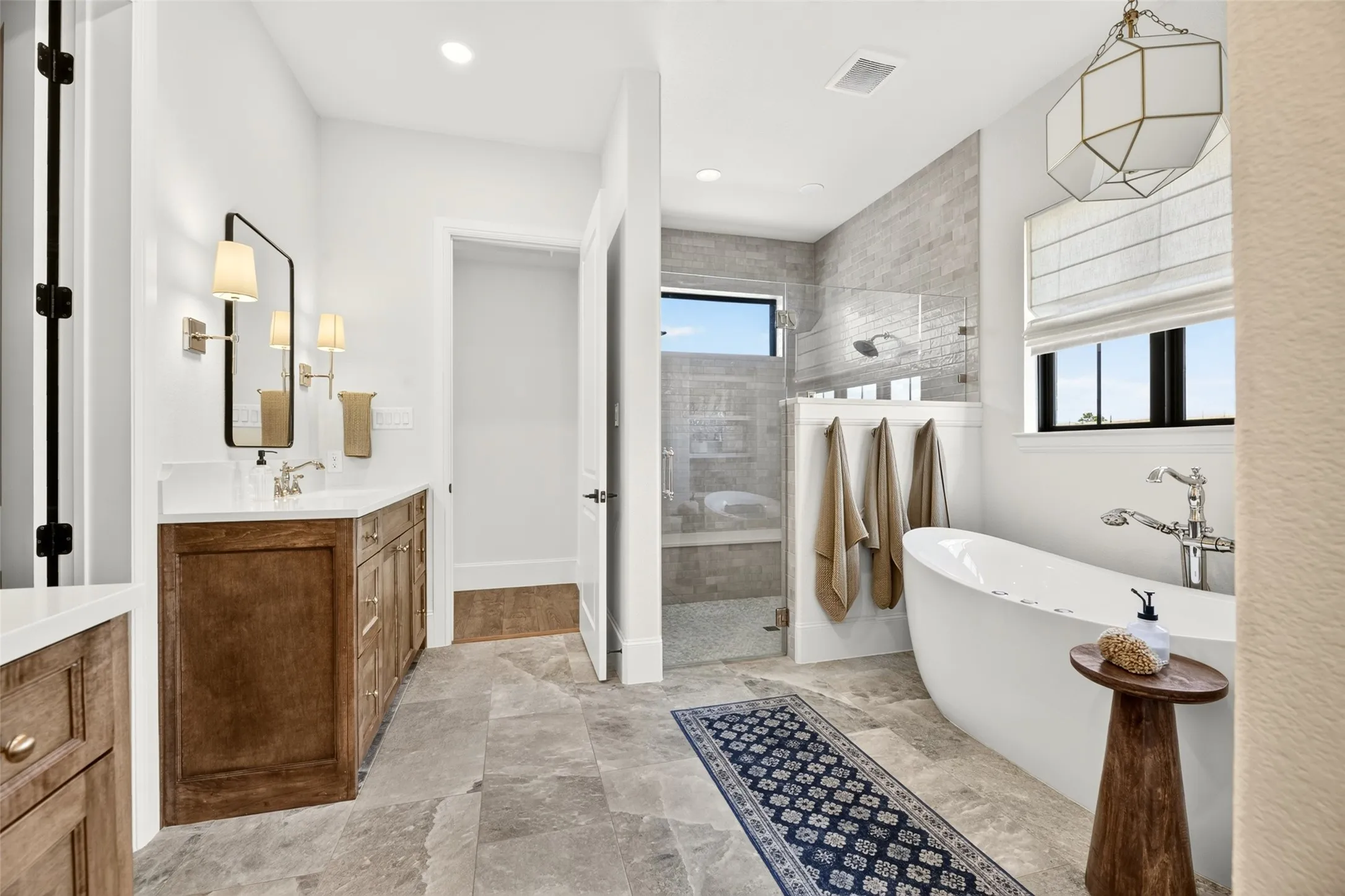 Freestanding soaking tub, walk-in shower with glass enclosure, wood-finish vanity with countertop, wall-mounted light fixtures, and large format tile flooring