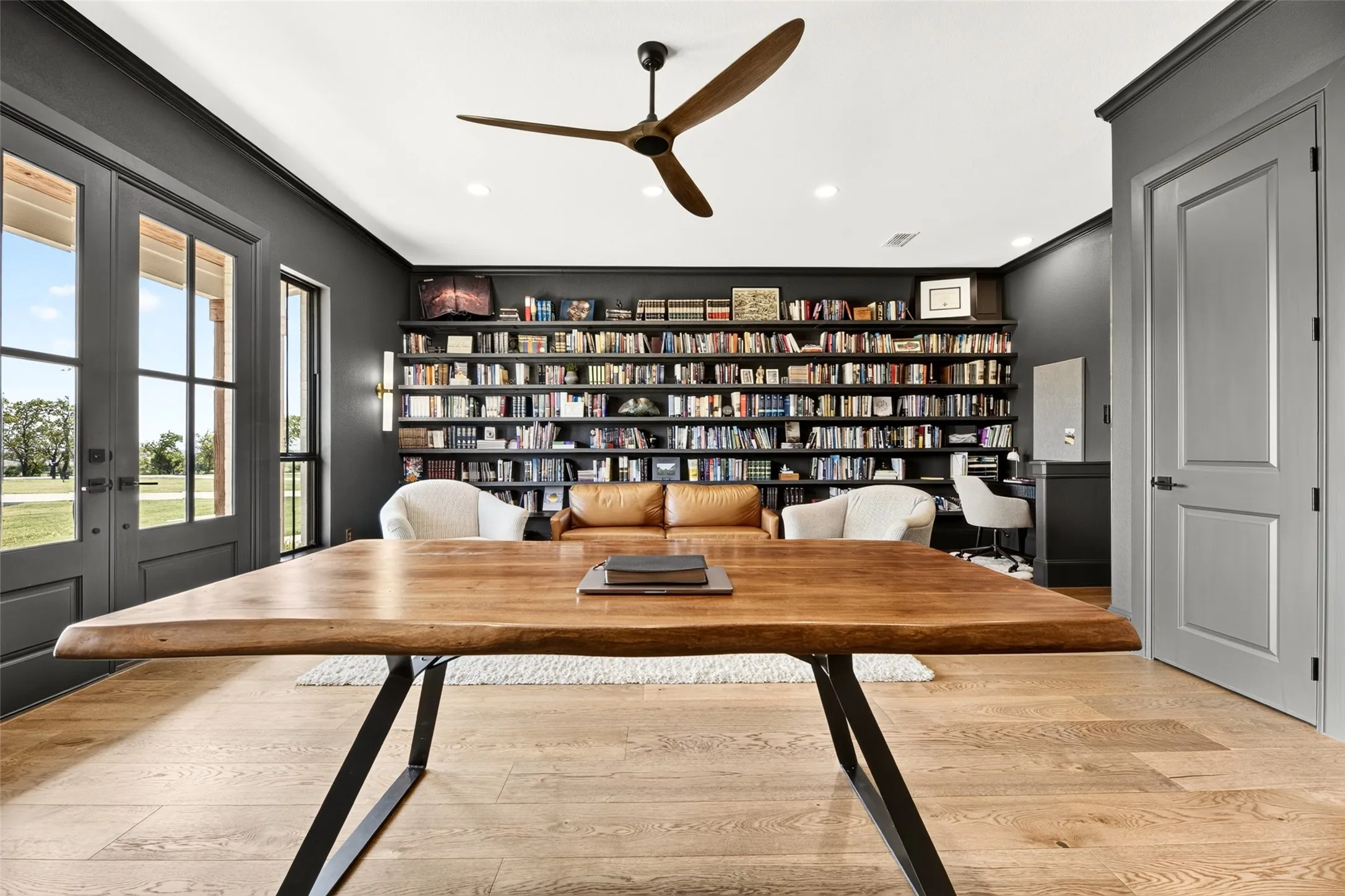 Wood-finish flooring with a natural grain, full-height built-in shelving, glass-paneled double doors, and a ceiling fan with wood-tone blades