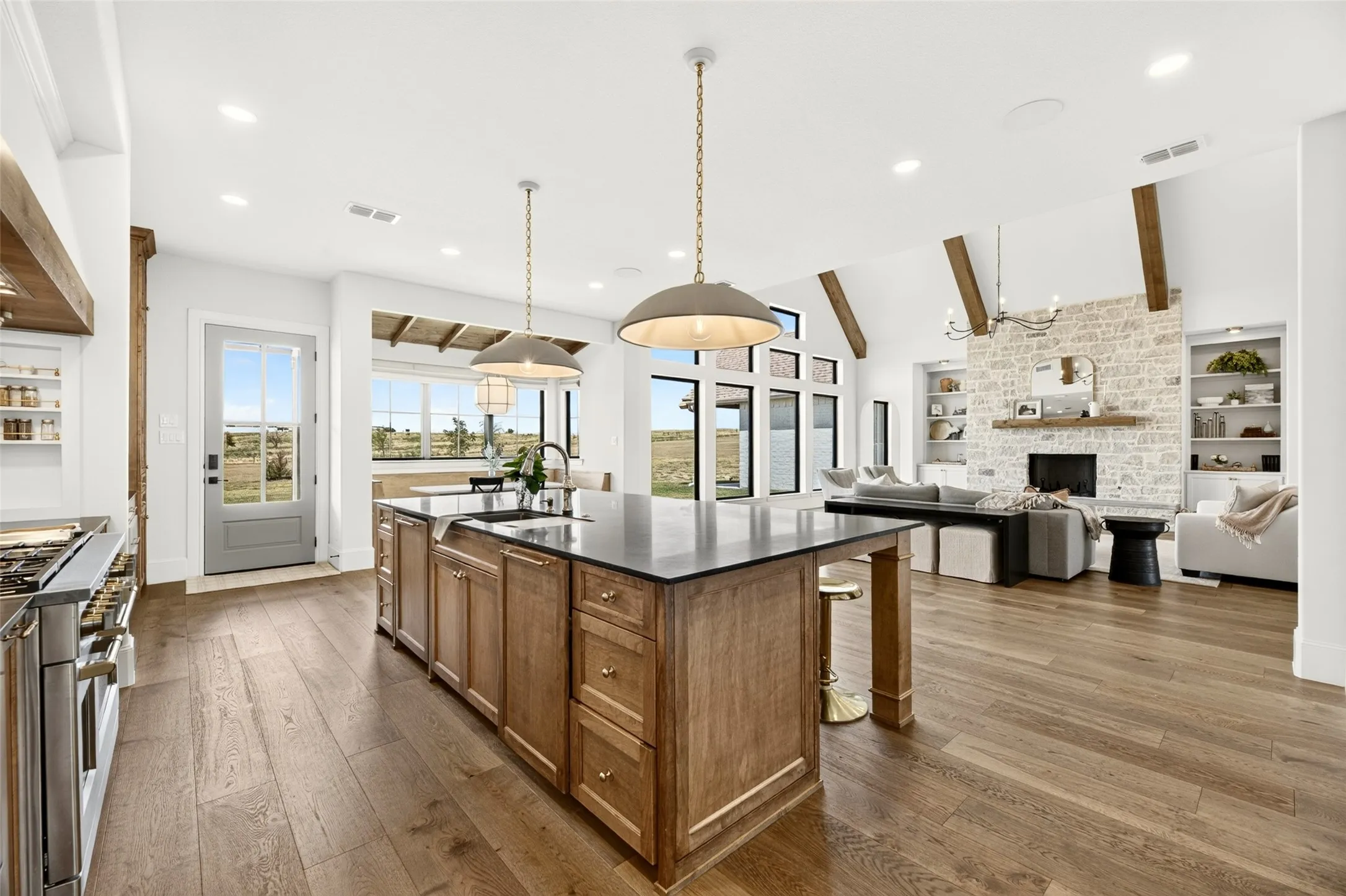 Expansive wood-finish flooring throughout, kitchen island with a dark countertop and pendant lighting, exposed ceiling beams, a stone-clad fireplace, and built-in shelving