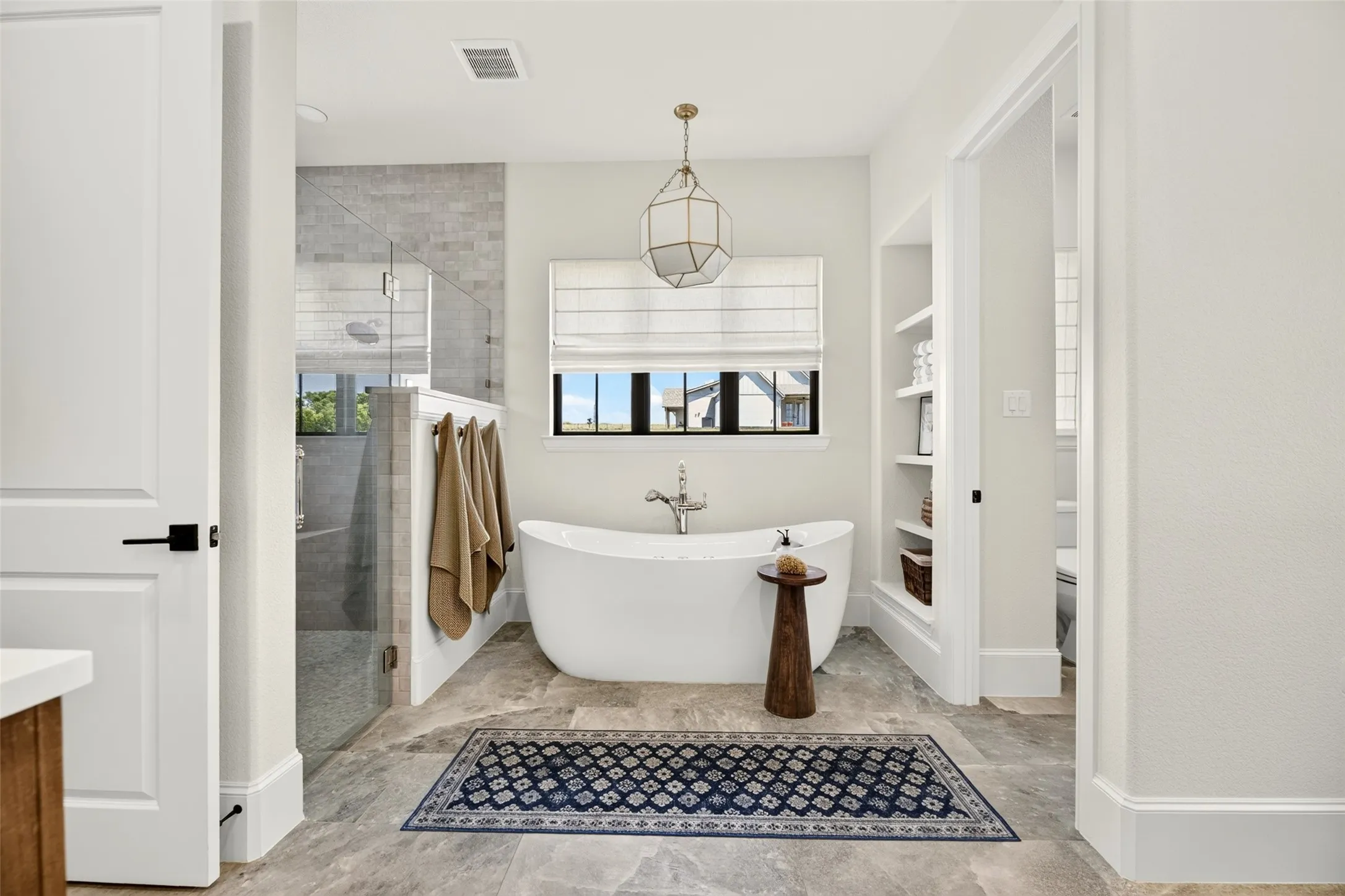Freestanding soaking tub, walk-in shower with glass enclosure, tiled shower surround, large window with Roman shade, and built-in shelving