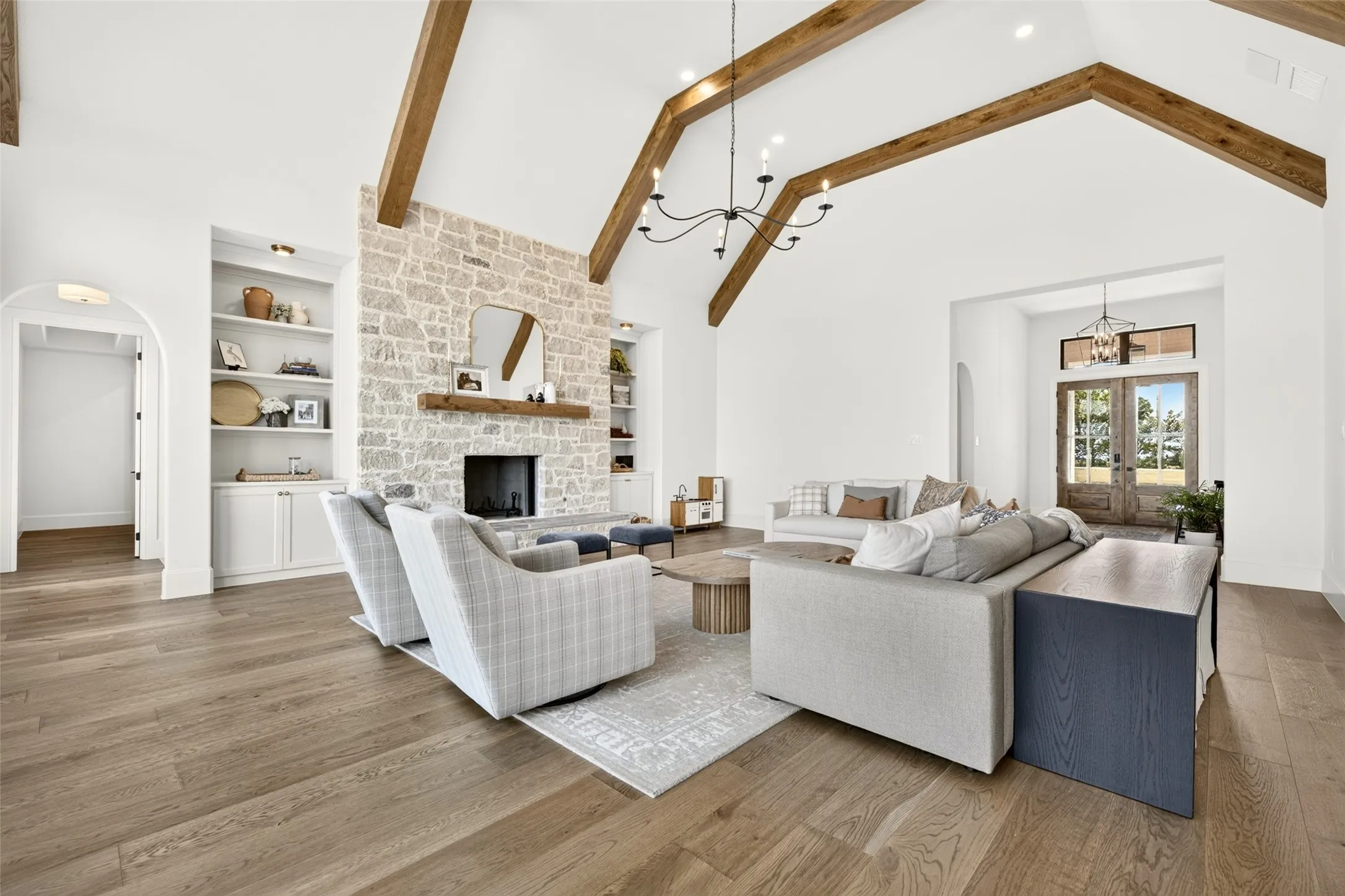 Vaulted ceilings with exposed wood beams, a stone-clad fireplace with a wood mantel, built-in shelving, wood-finish flooring, and double entry doors with transom window