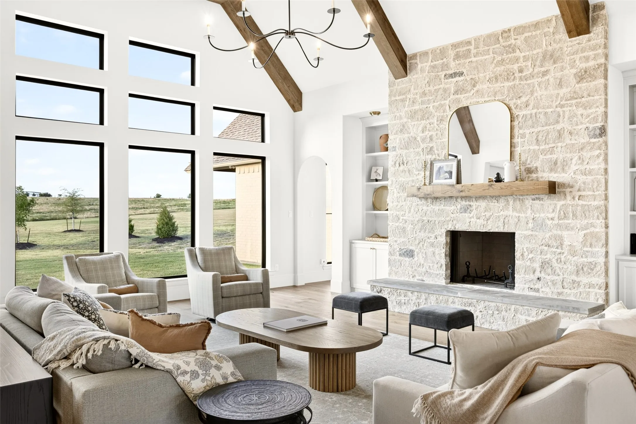 Vaulted ceiling with exposed wood beams, stone-clad fireplace with wood mantel, built-in shelving, large black-framed windows, and light wood-finish flooring