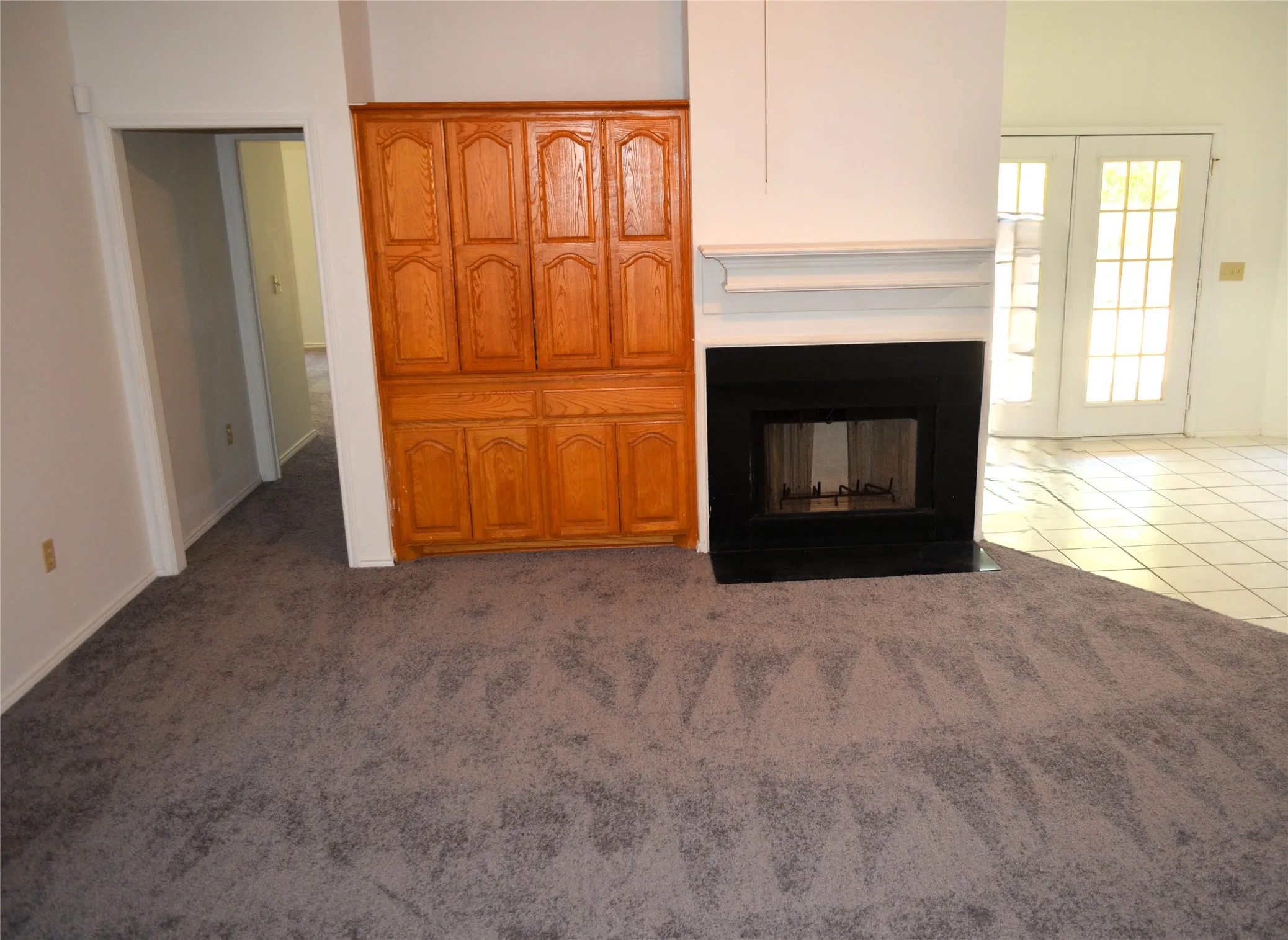 Carpeted flooring, built-in wooden cabinetry, fireplace with a mantel, and double French doors opening to tile flooring