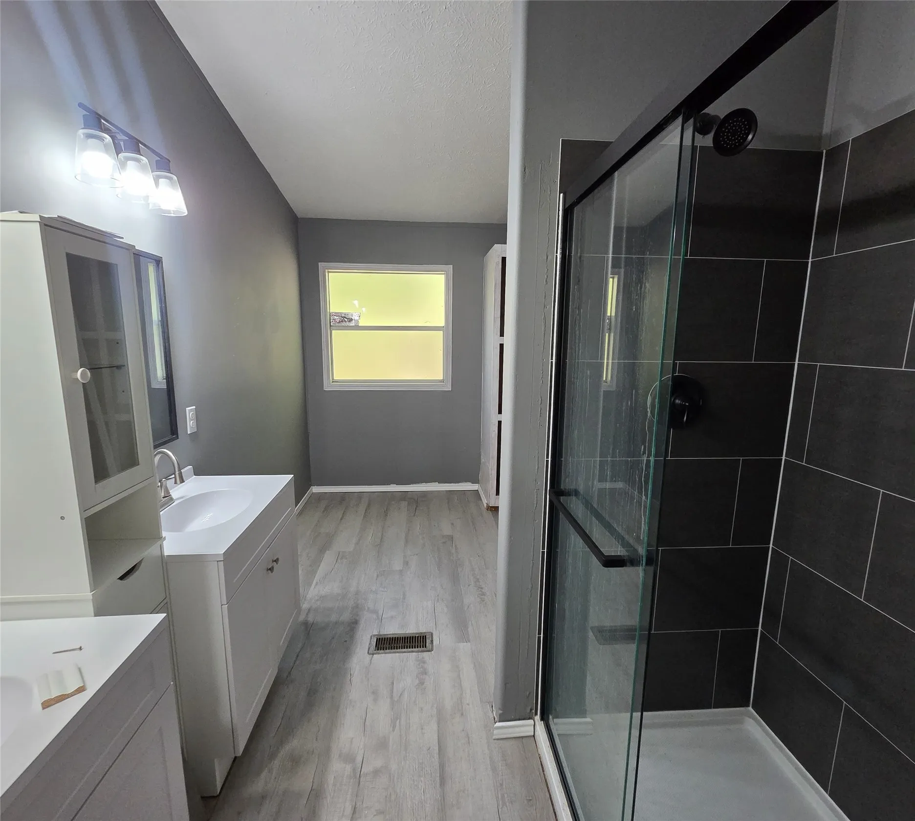 Dual vanity sinks, wood-looking finish flooring, dark tiled shower, and a glass-enclosed shower