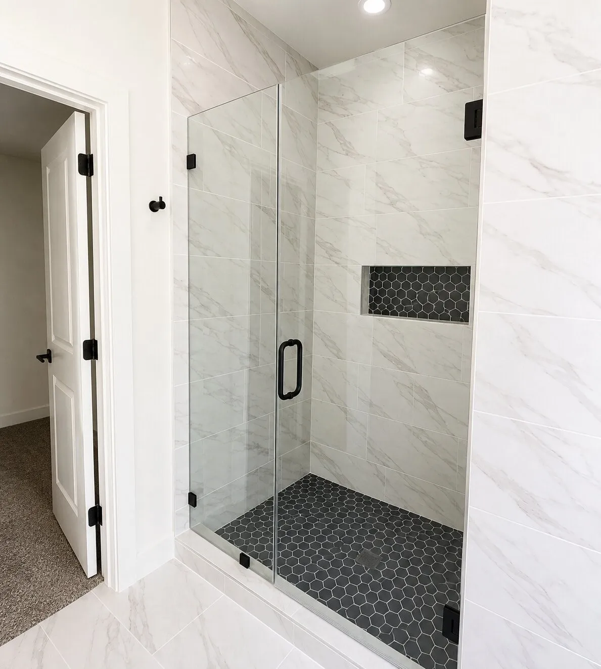 Glass-enclosed shower with a black hexagonal tile floor, a recessed niche with hexagonal tile, and large format white tile walls