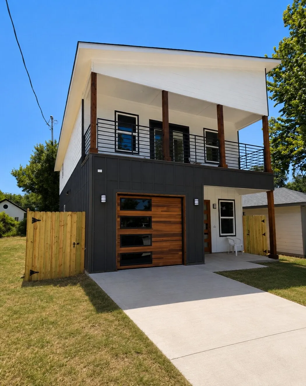 Modern exterior with contrasting dark and white siding, wood garage door with frosted glass panels, upper-level balcony with horizontal bar railing, wood columns, and clear concrete driveway