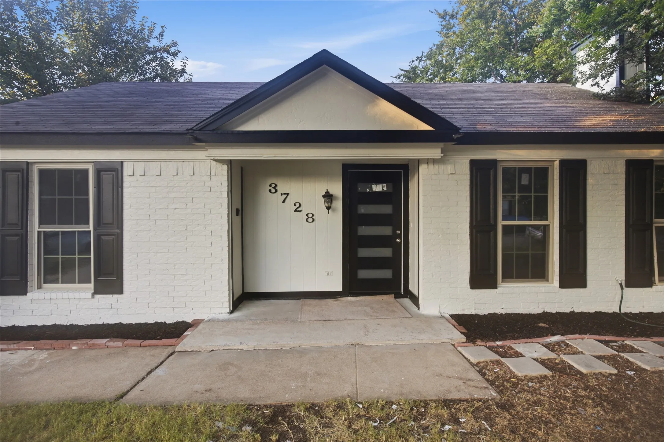 White brick exterior with dark shutters, a dark roof, and an asphalt driveway