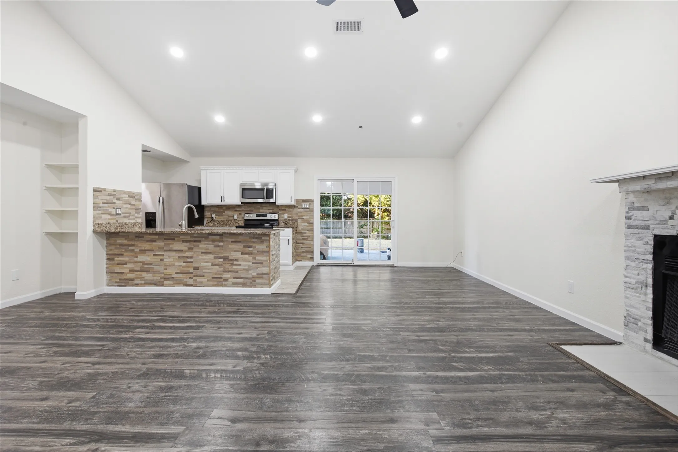 Vaulted ceiling with recessed lighting, wood-finish flooring, stone-clad fireplace, kitchen with white cabinetry and stainless steel appliances, and a sliding glass door