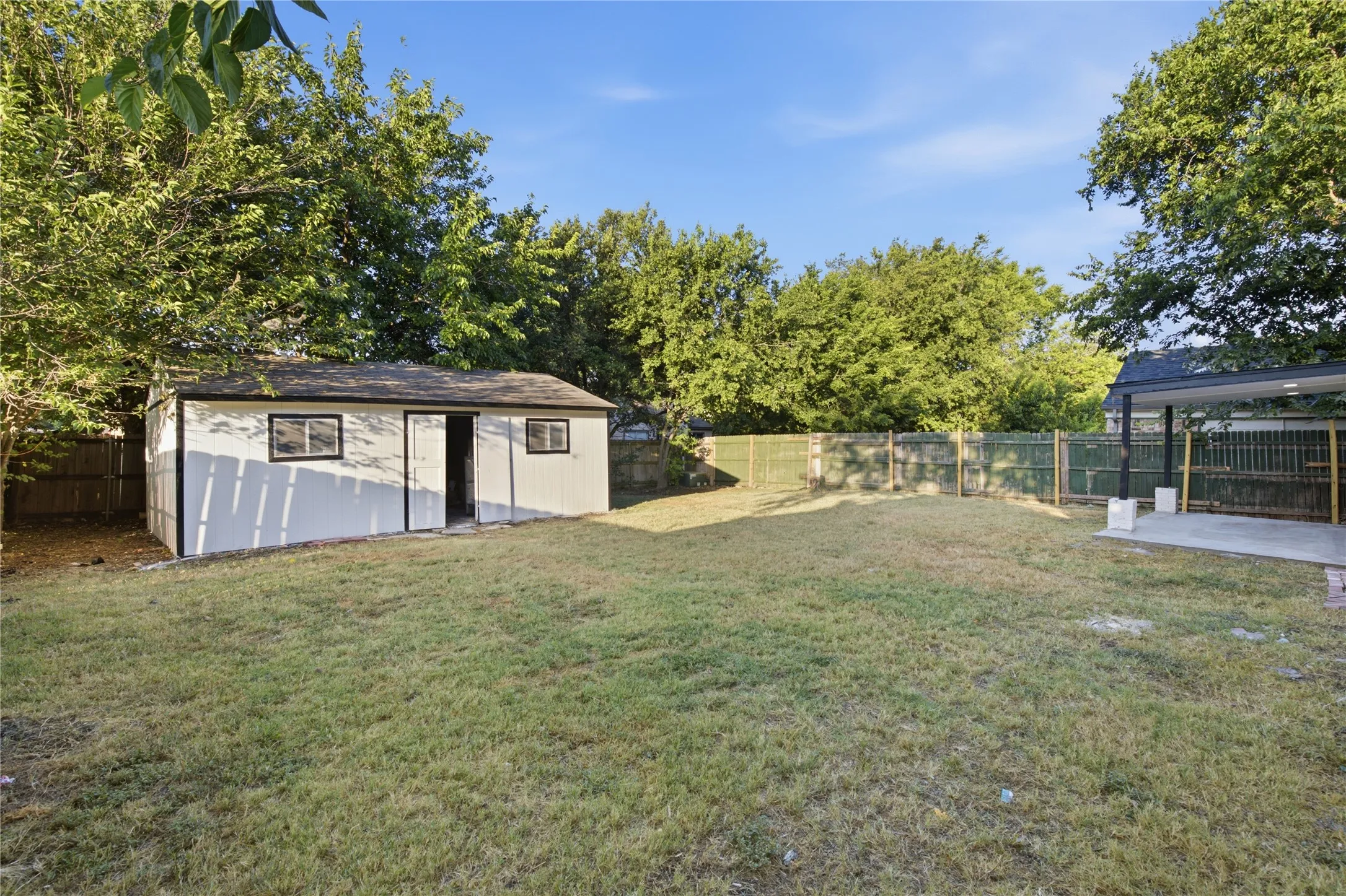 Expansive yard featuring a detached storage building with multiple windows, a paved patio, and mature trees providing natural privacy