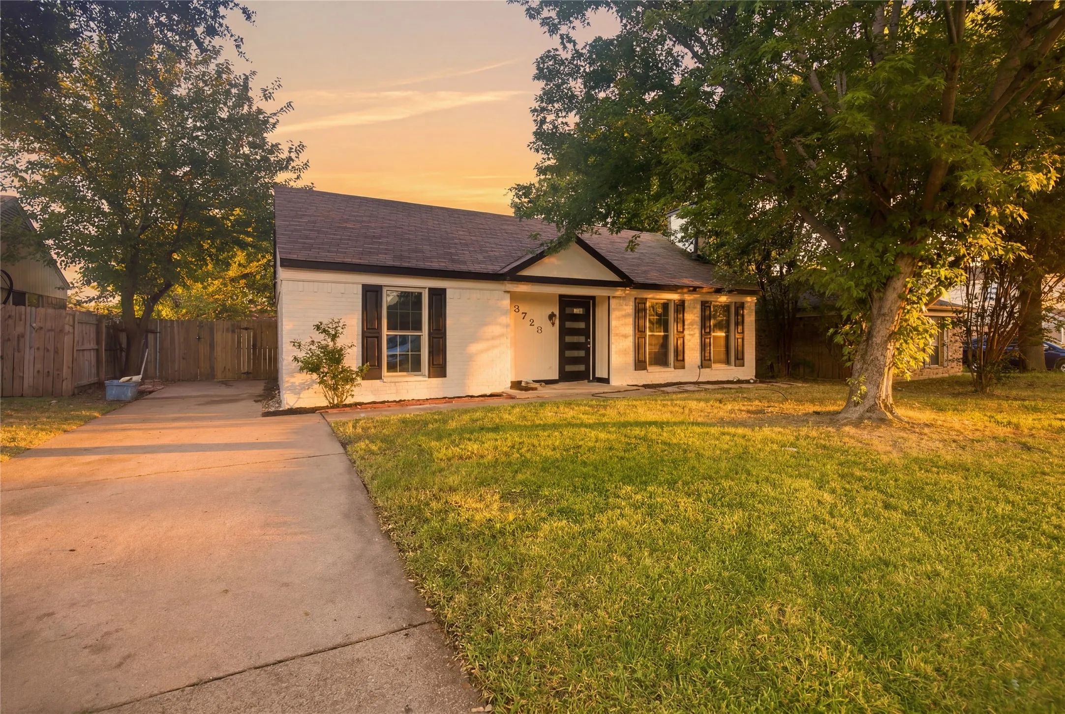White brick exterior, dark composition shingle roof, dark wood shutters, front yard with mature trees, and a concrete driveway