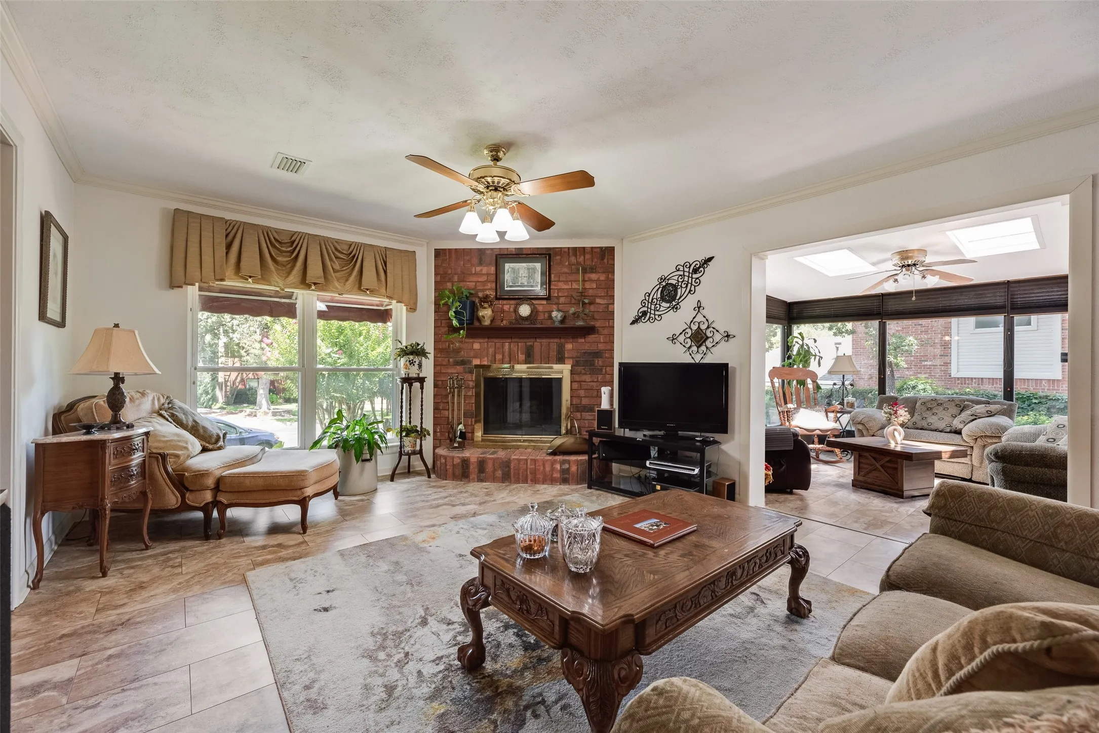 Corner brick fireplace with hearth, ceiling fan with integrated lighting, tile flooring, and a sunroom with skylights